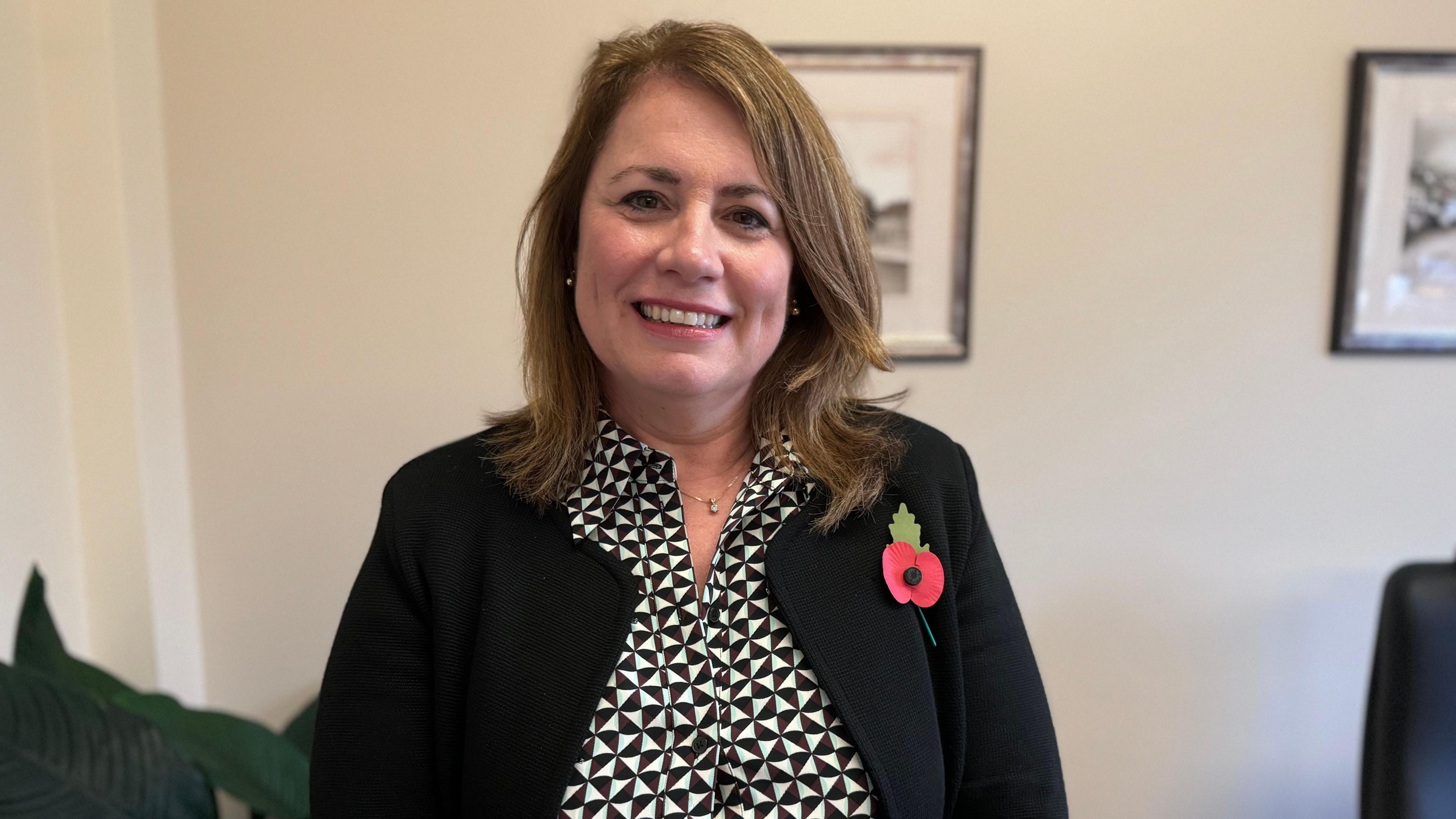 Miss Evans smiles while wearing a black and white top with a black jacket. She has brown shoulder length hair. A red poppy is clipped to her jacket.