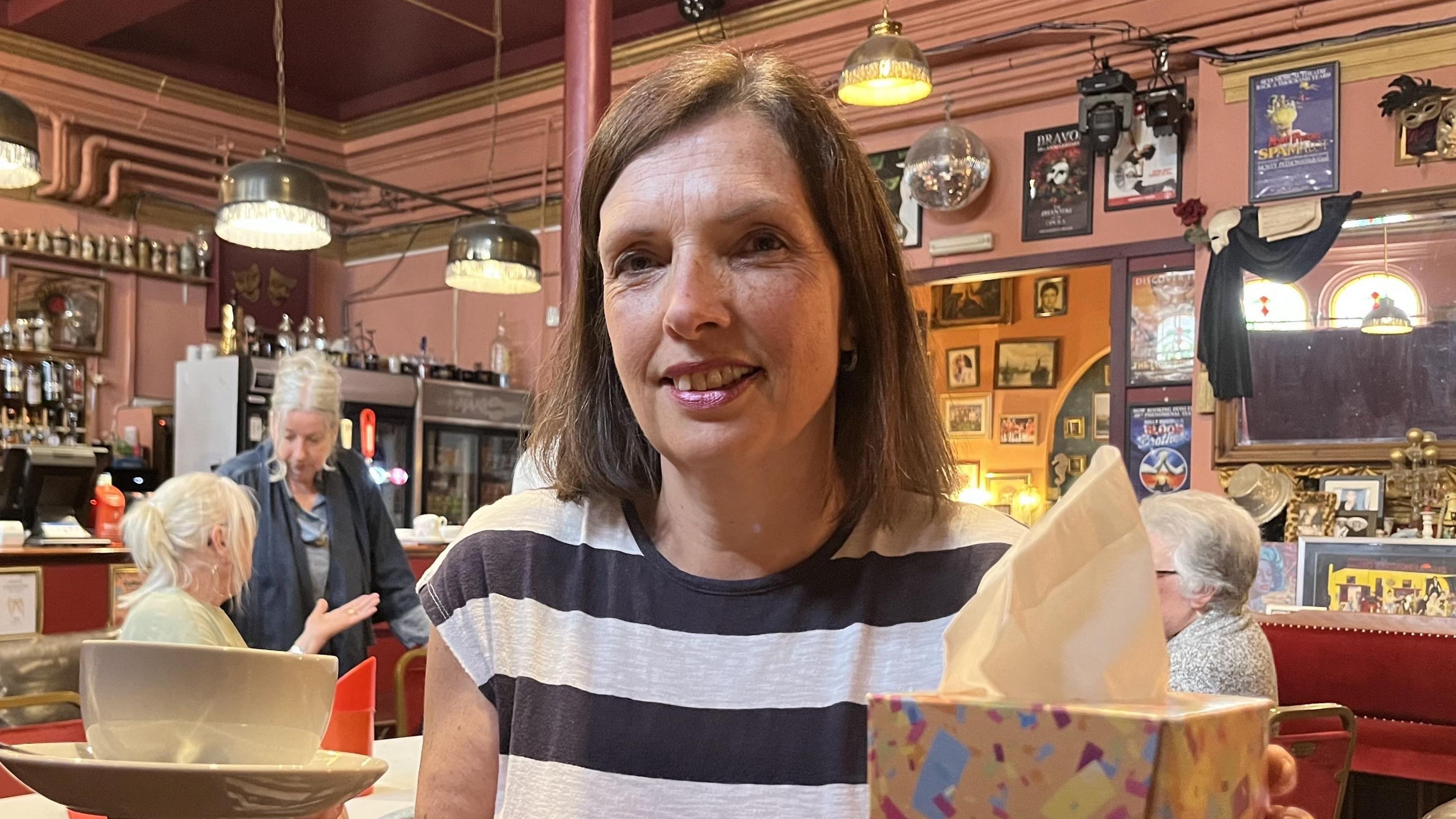 Thomasina Scott-Greenard, wearing a blue and white short-sleeve hooped top, sits in a cafe with several people behind her. She has dark, shoulder-length hair and is holding a coffee cup and a box of tissues.