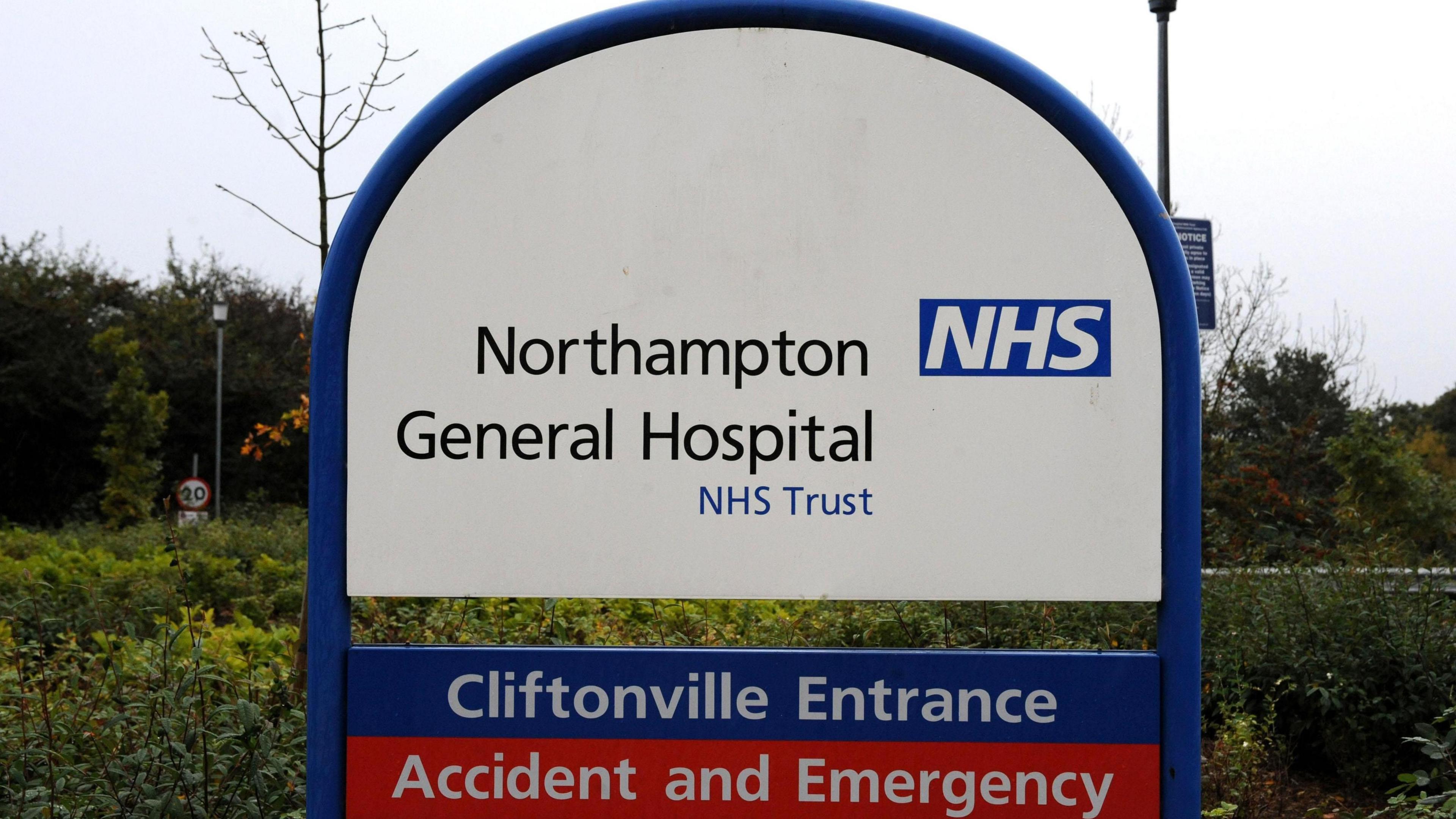 A front on photo of a hospital sign. The sign is blue white and red and it in a semi circle shape. The top of the sign is white and says Northampton General hospital.