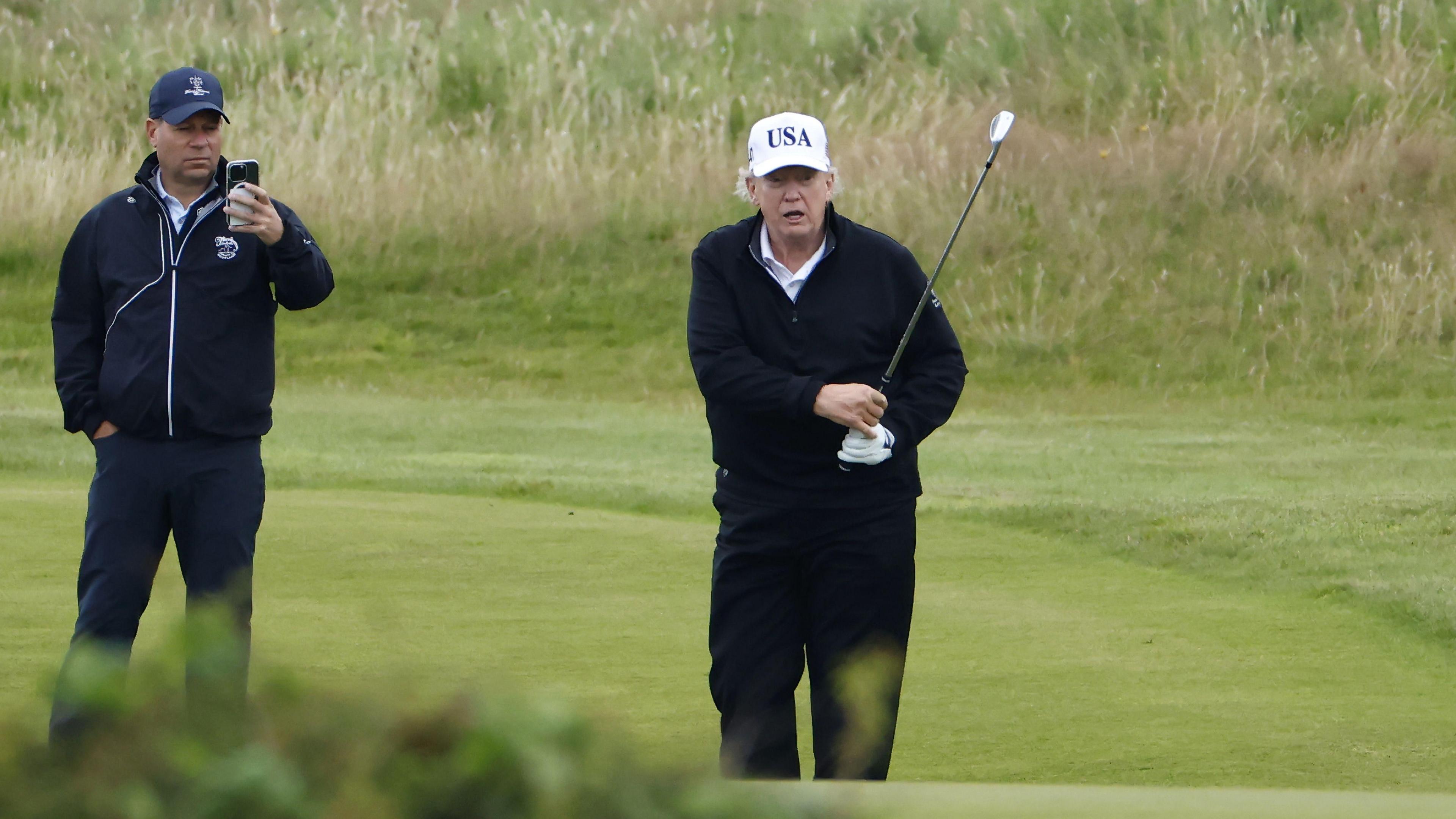 Trump completes his golf swing and follows looks as his ball heads off. A man in golfwear and hat and filming on a phone is behind him