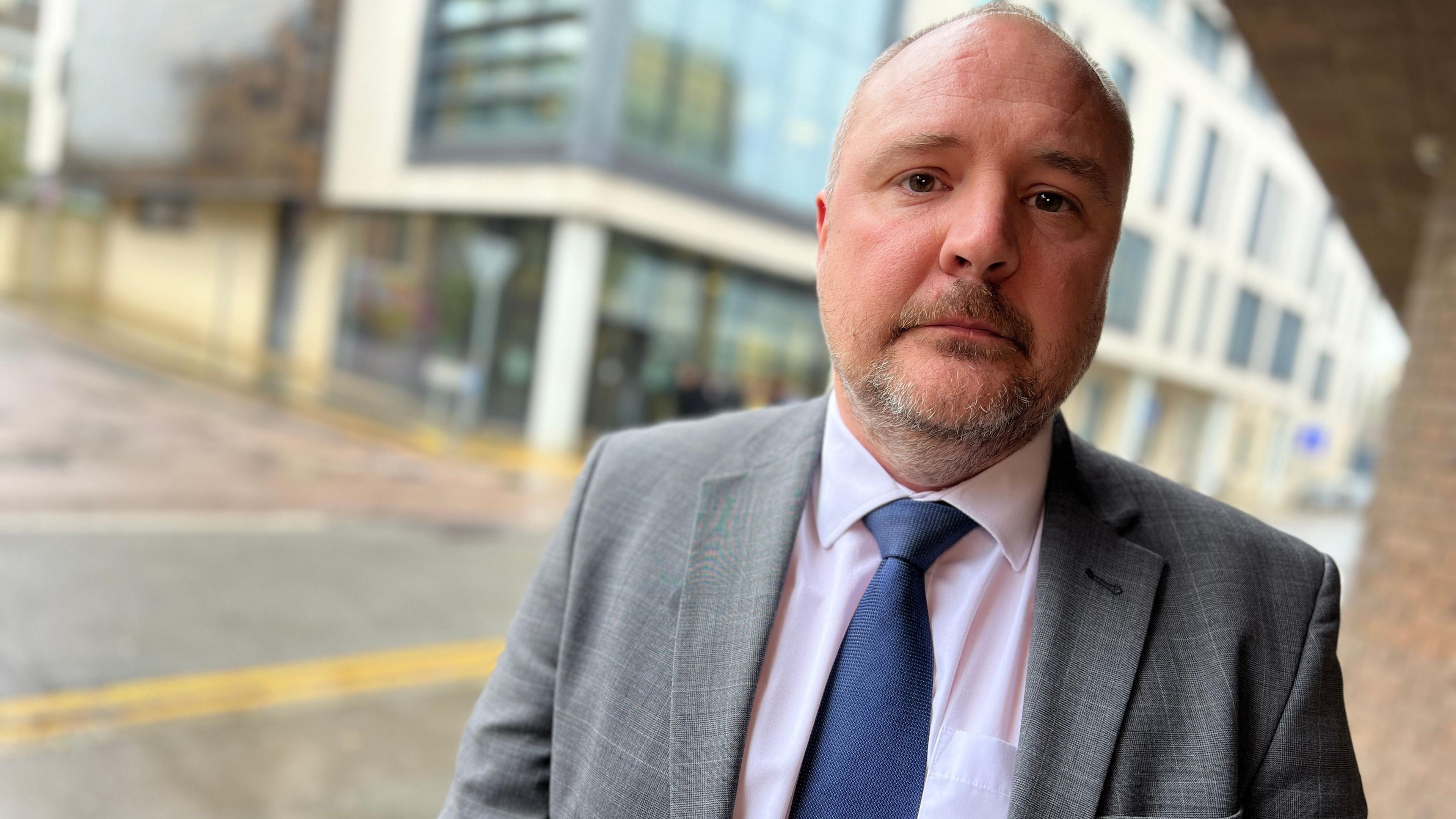 Rob Kirby standing outside Chelmsford Crown Court. He is wearing a grey suit jacket, pink shirt and navy tie. He is looking seriously at the camera.