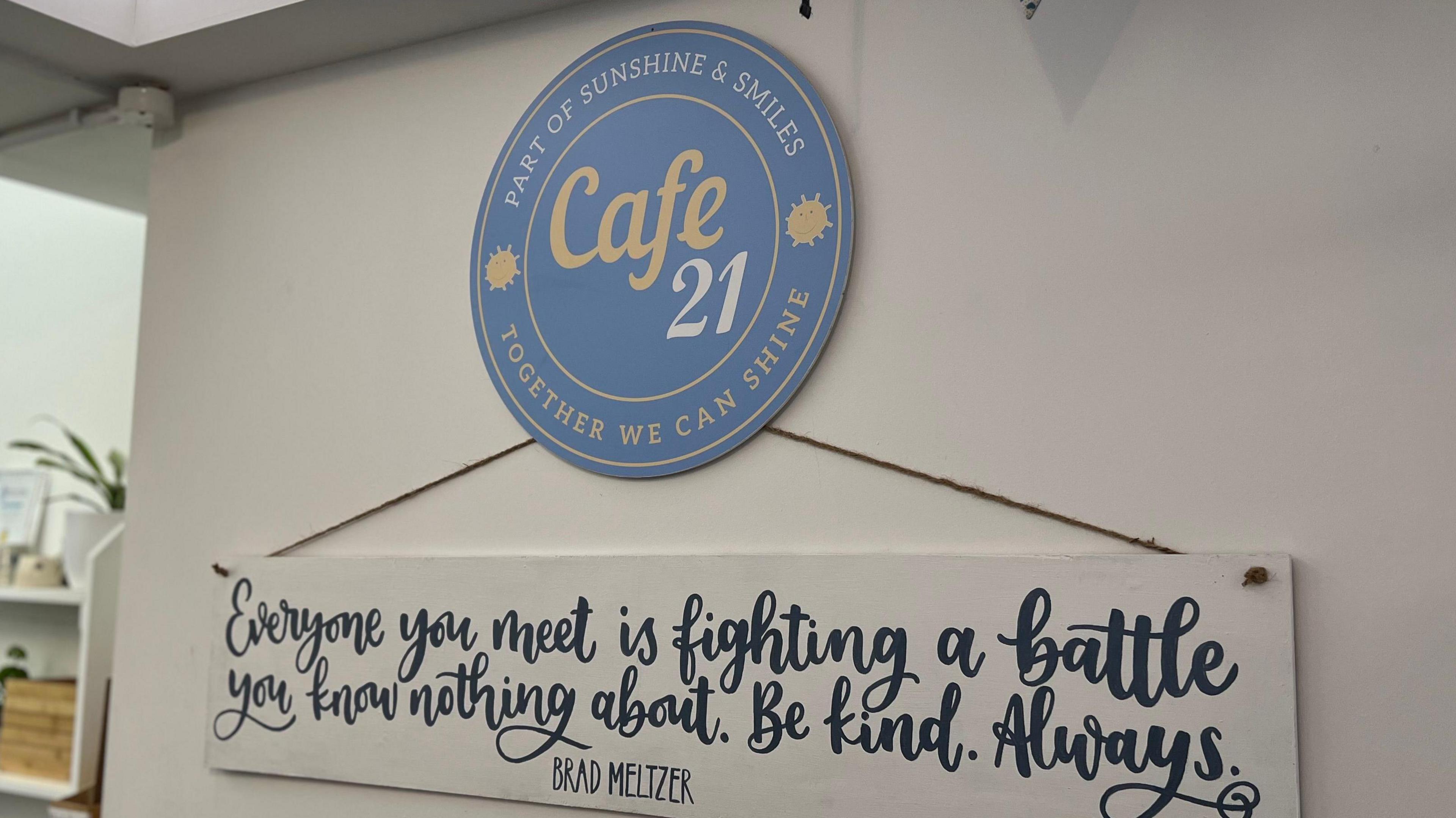 Picture of cafe 21's sign hanging on a wall and a quote saying: "Everyone you meet is fighting a battle you know nothing about. Be kind. Always."