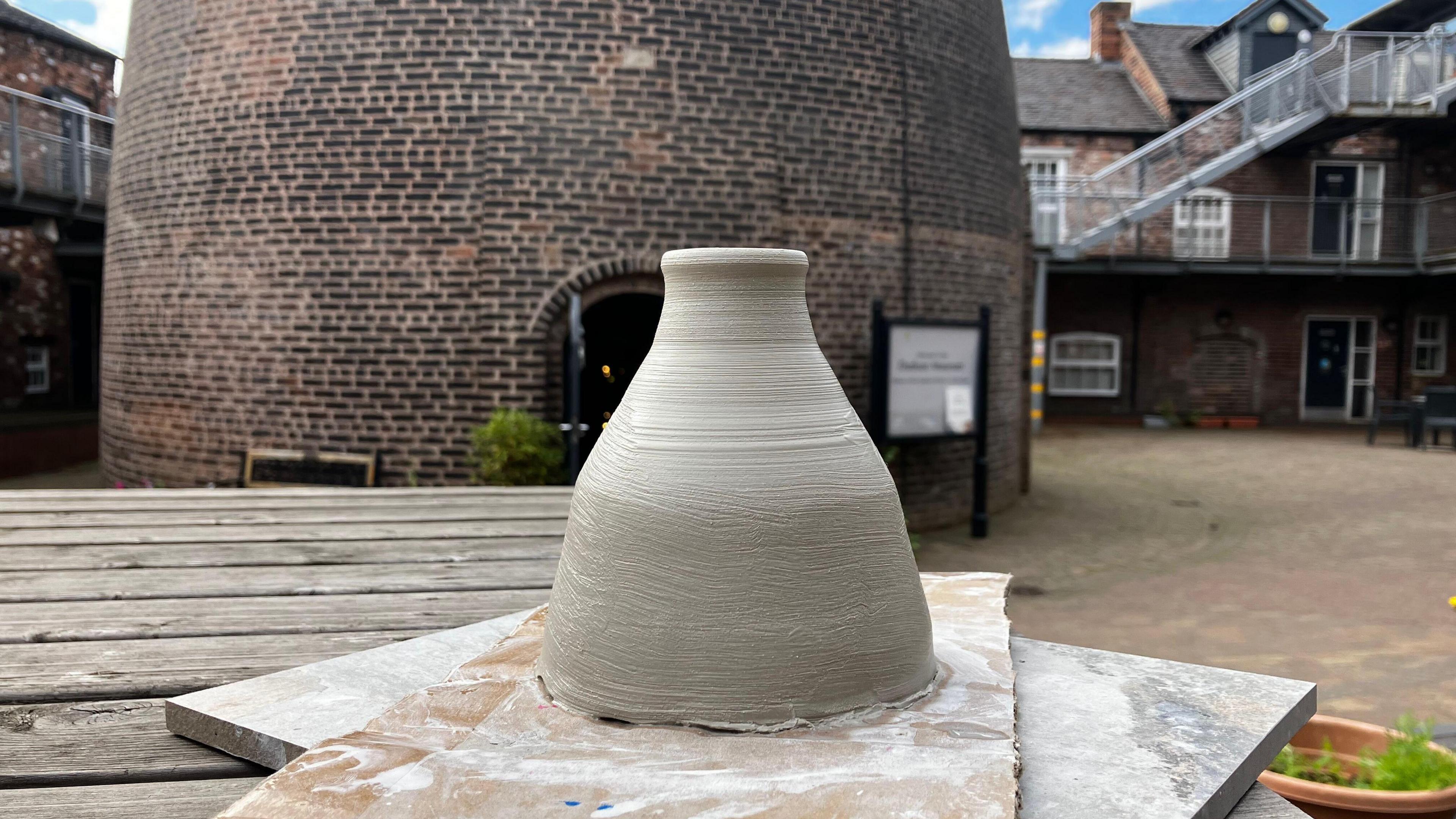 A miniature bottle kiln that has been freshly moulded out of clay. It has a wide base and a narrow neck and is on a wooden board and in front of a large bottle kiln, called Dudson Kiln.