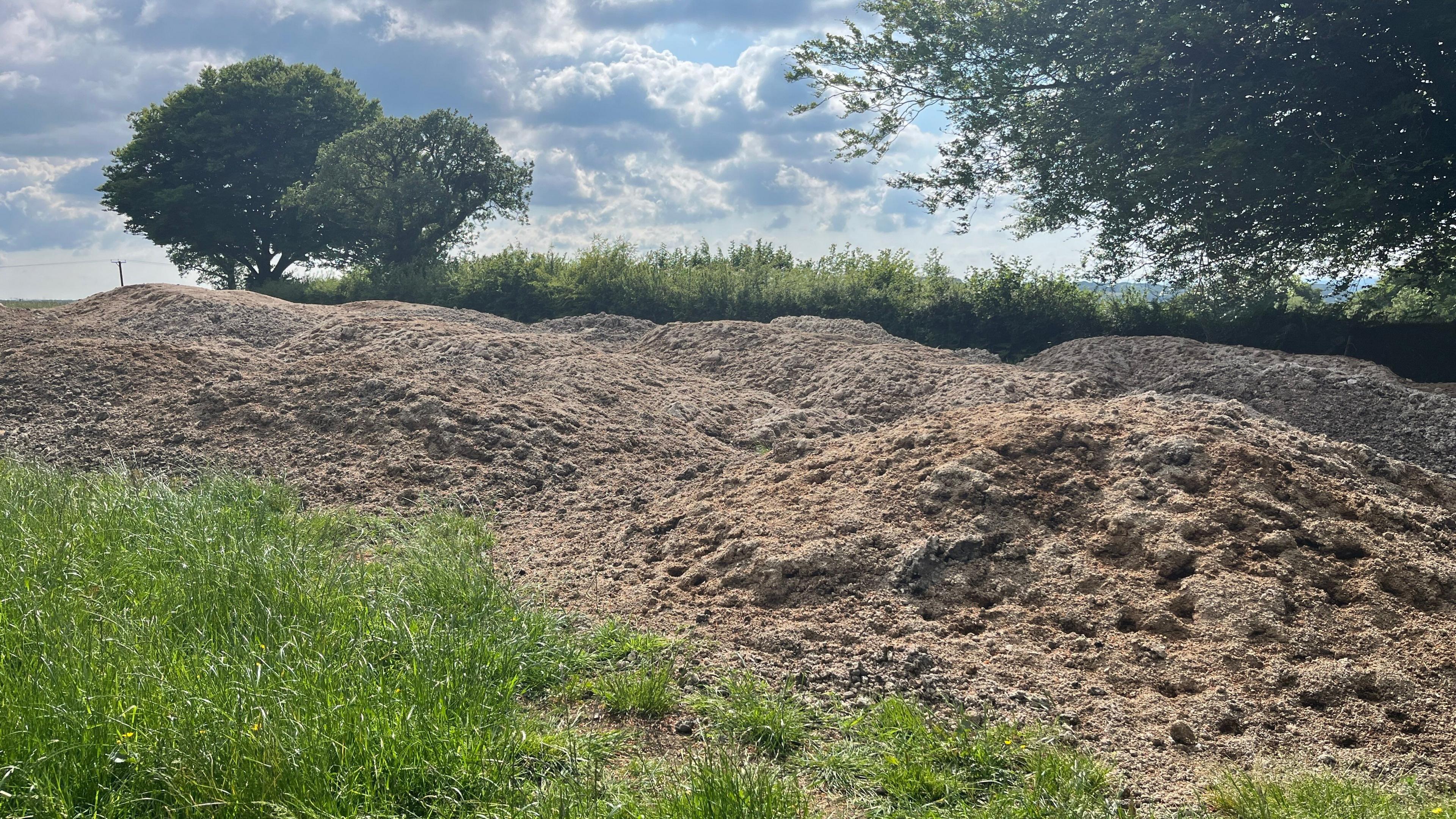 A large brown pile of sewage sludge in a field surrounded by grass and trees. 