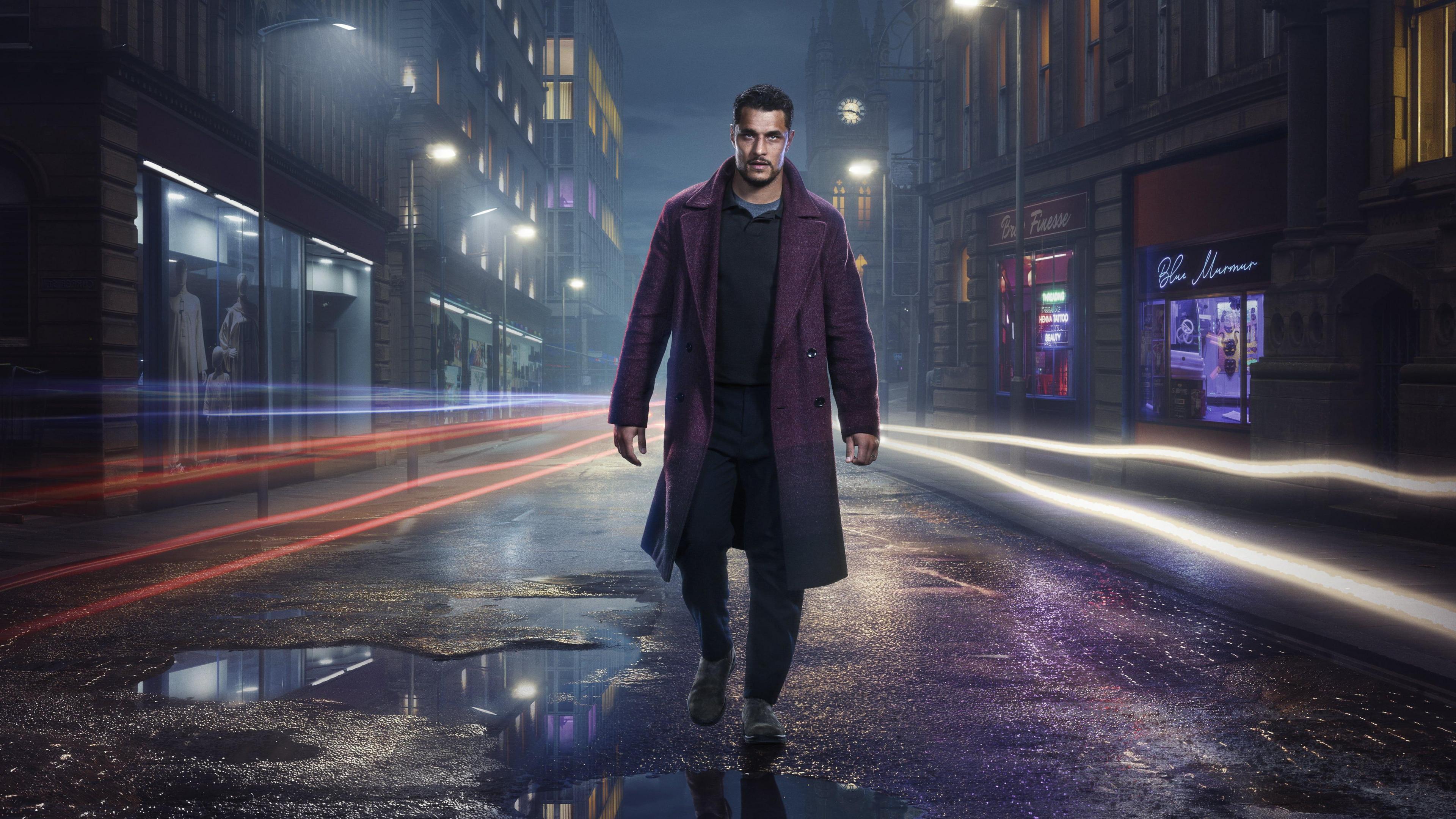 A man walks down a wet street in the dark in an artistic promo image. Bolts of light run alongside him on both sides. He is wearing a burgundy coat and black top and trousers.