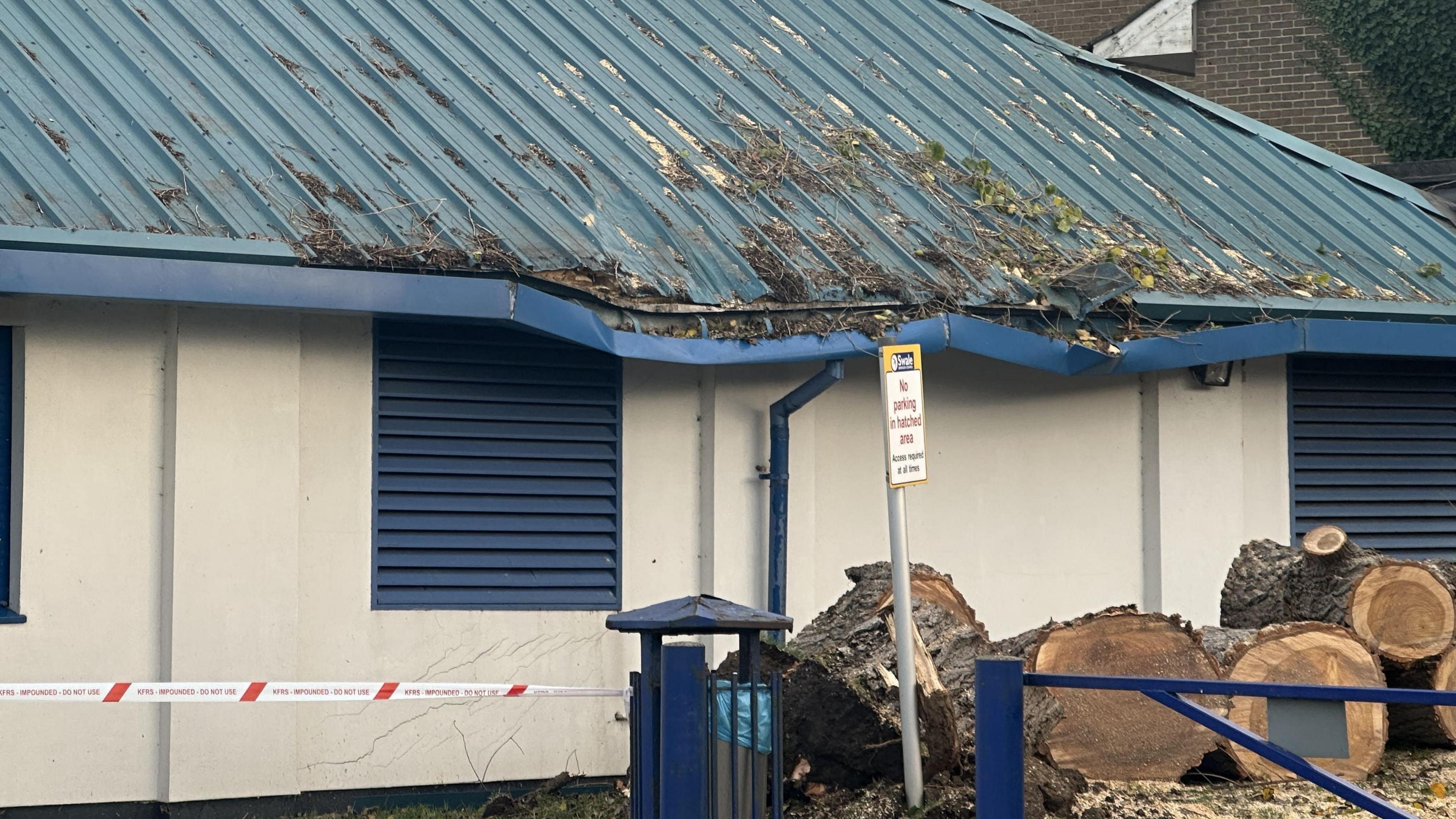 A cut-up tree at the base of a building which has a damaged blue roof.