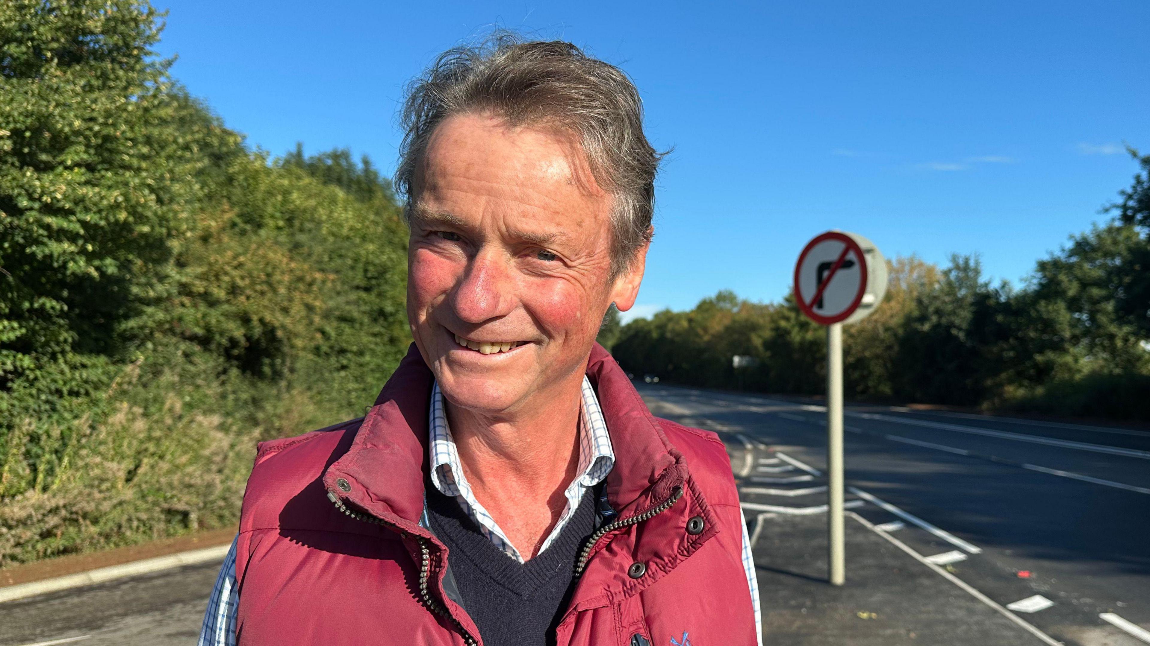 Gideon Sumption. He has short grey hair and is wearing a checked shirt, a navy blue jumper and a red gilet. He is pictured near a road junction. He is looking at the camera and smiling.