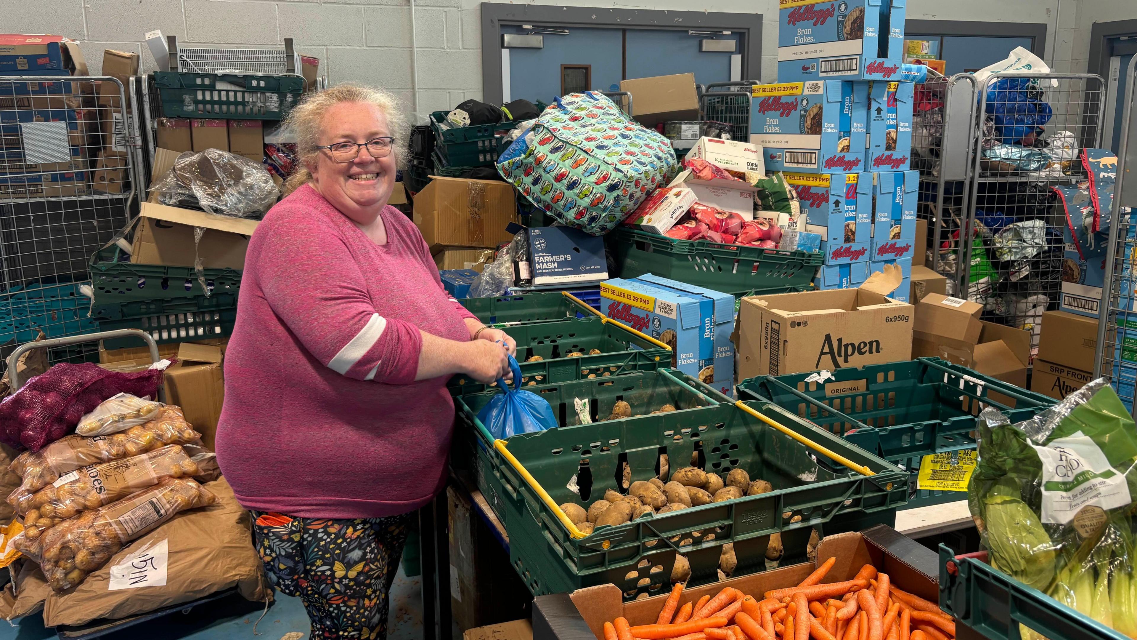 A smiling Alainea Stark wearing a dark pink jumper and leggings stands in a store room next to a table with crates of vegetables including carrots and potatoes, with other food goods stacked behind her