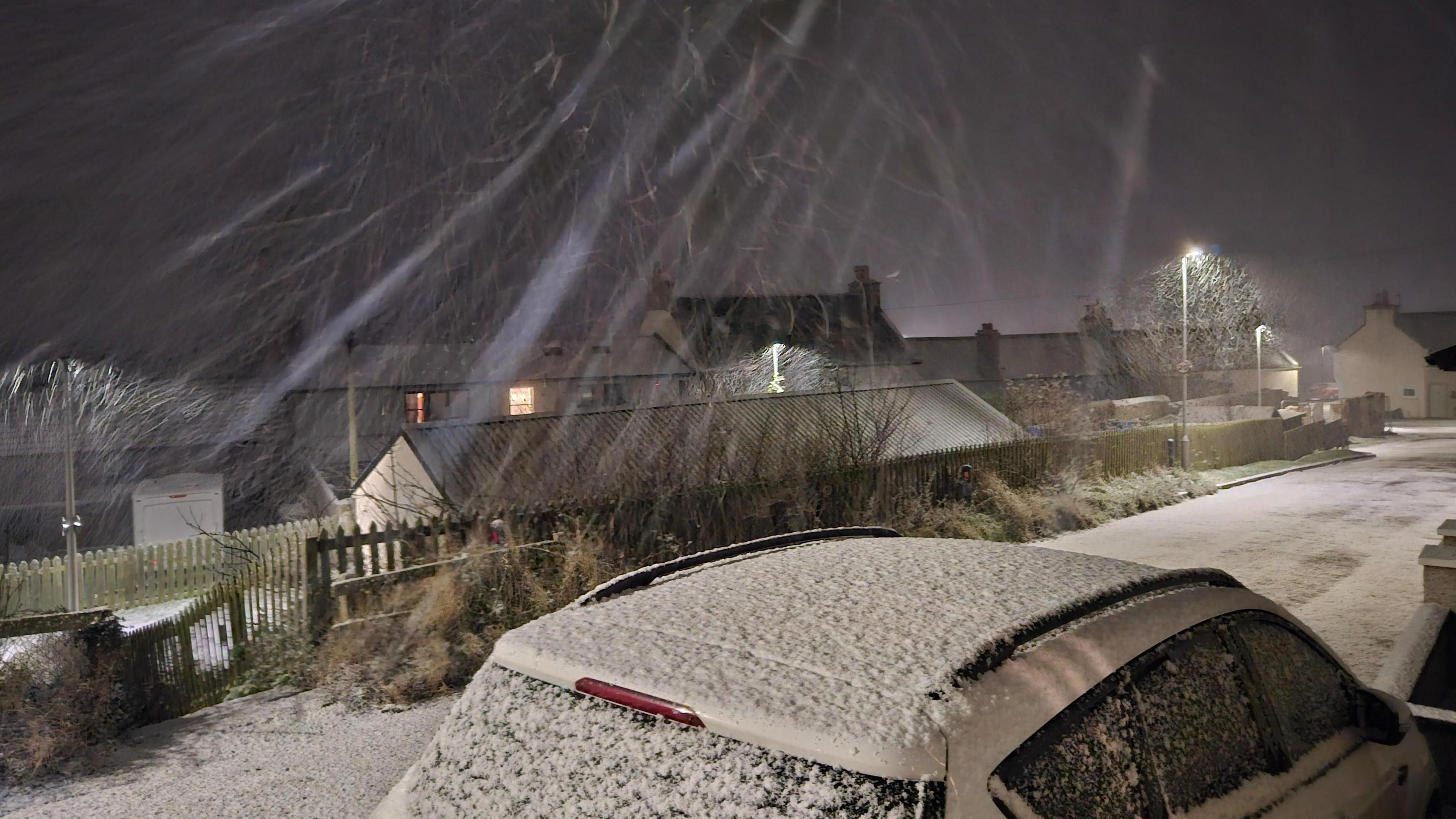 Snow lands on top of a white car. The streets are also covered in snow and is falling from a dark sky.