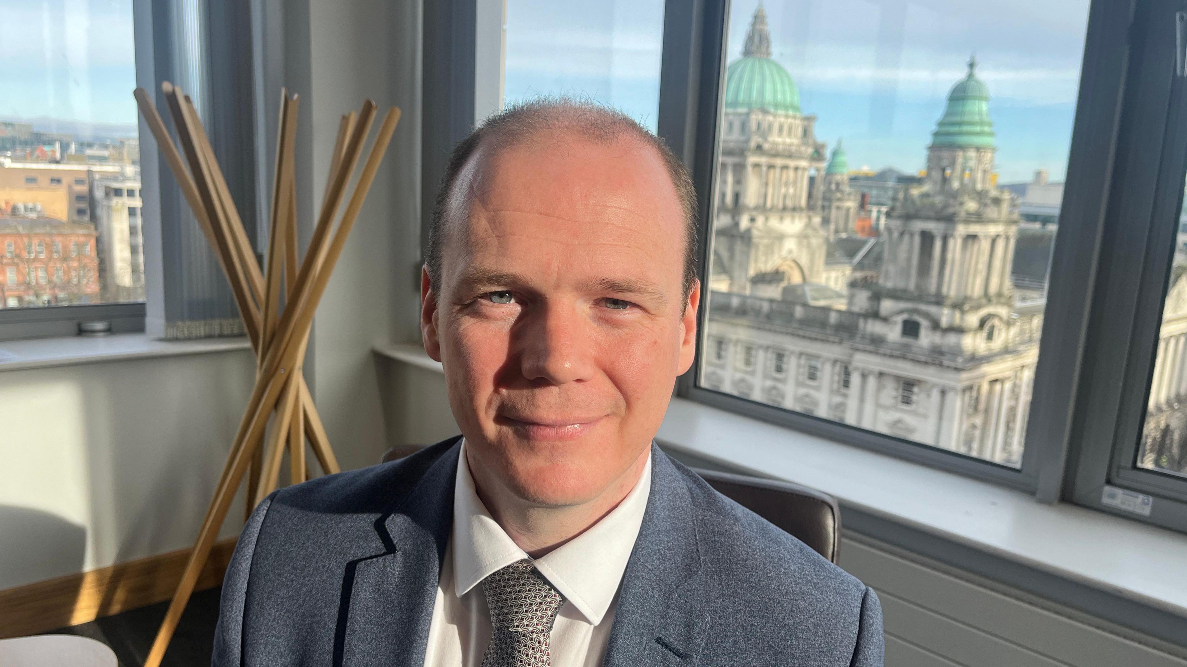 Communities minister Gordon Lyons sits in front of a series of windows out of which the belfast skyline, including city hall, can be seen. He is balding, wearinhg a grey suit jacket , white shirt and grey tie. 