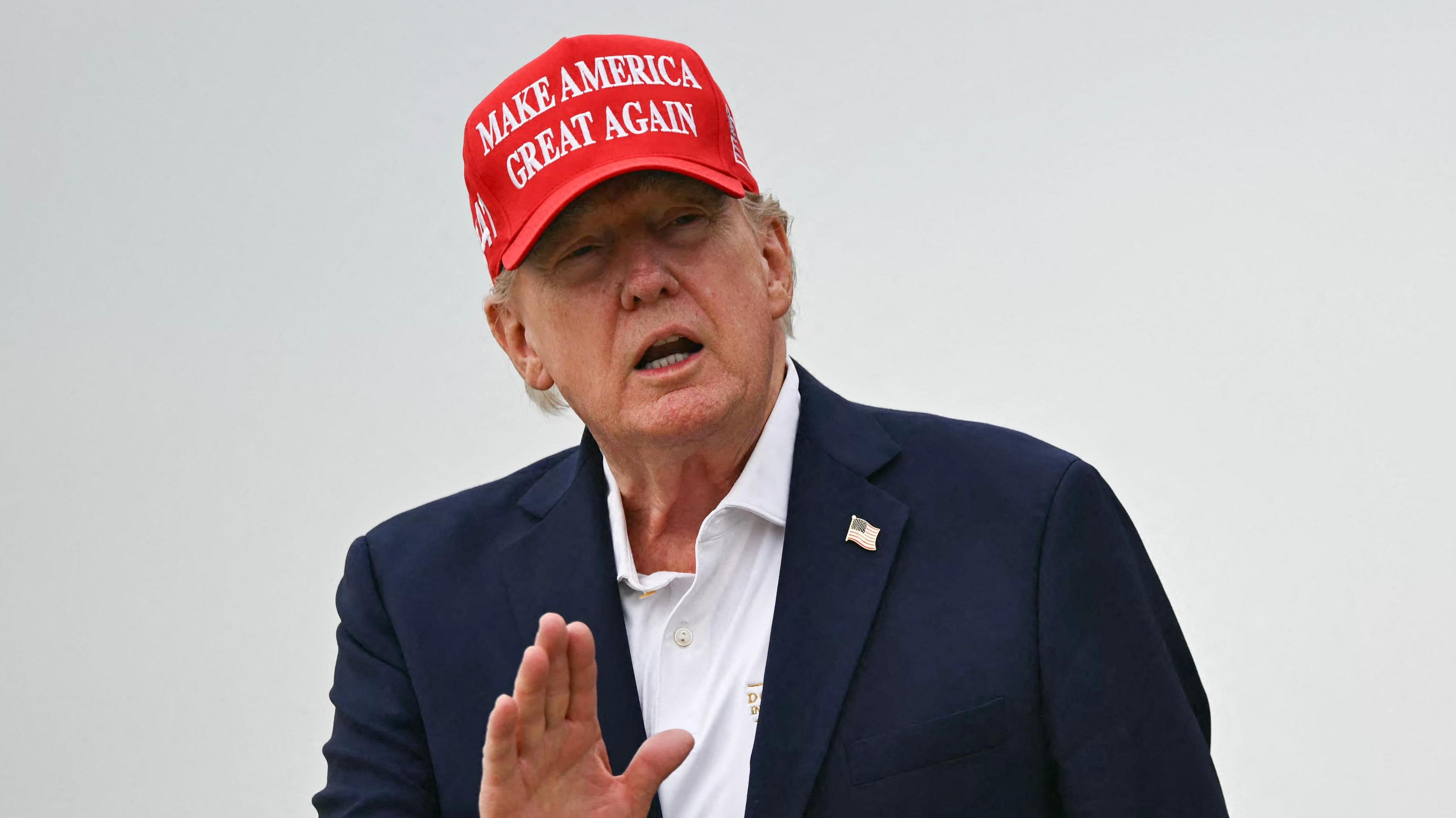 A close up shot of President Donald Trump speaking to outside with his hand raised. He is wearing a "make America great again" cap