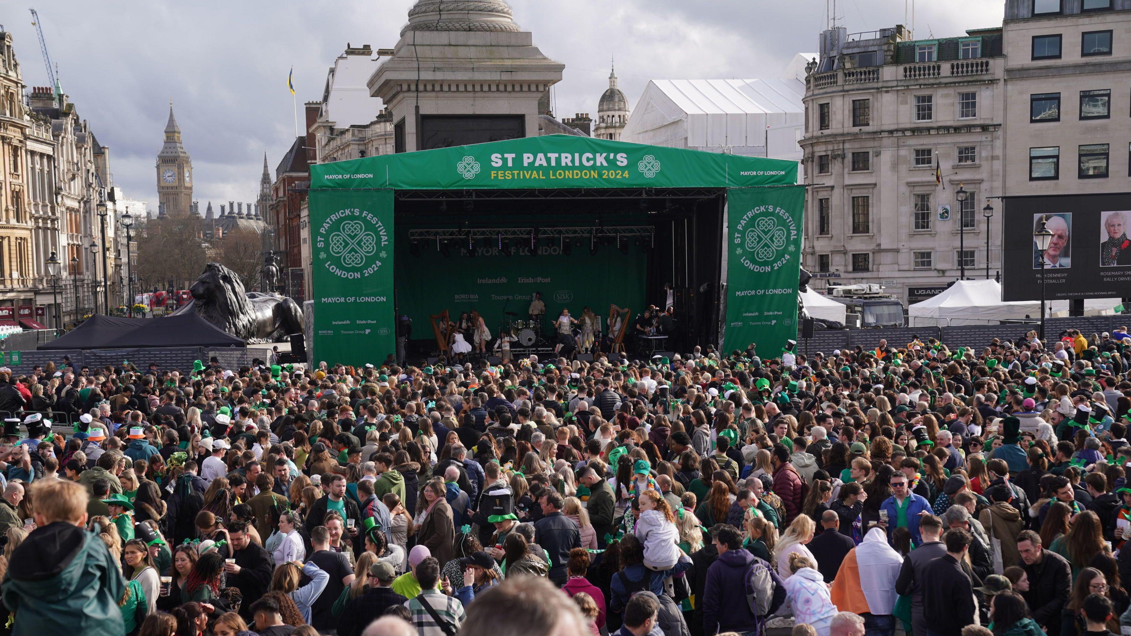 An image showing crowds of people looking towards a stage in Trafalgar Square covered in green tarpaulin saying 'St Patrick's Festival London 2024'