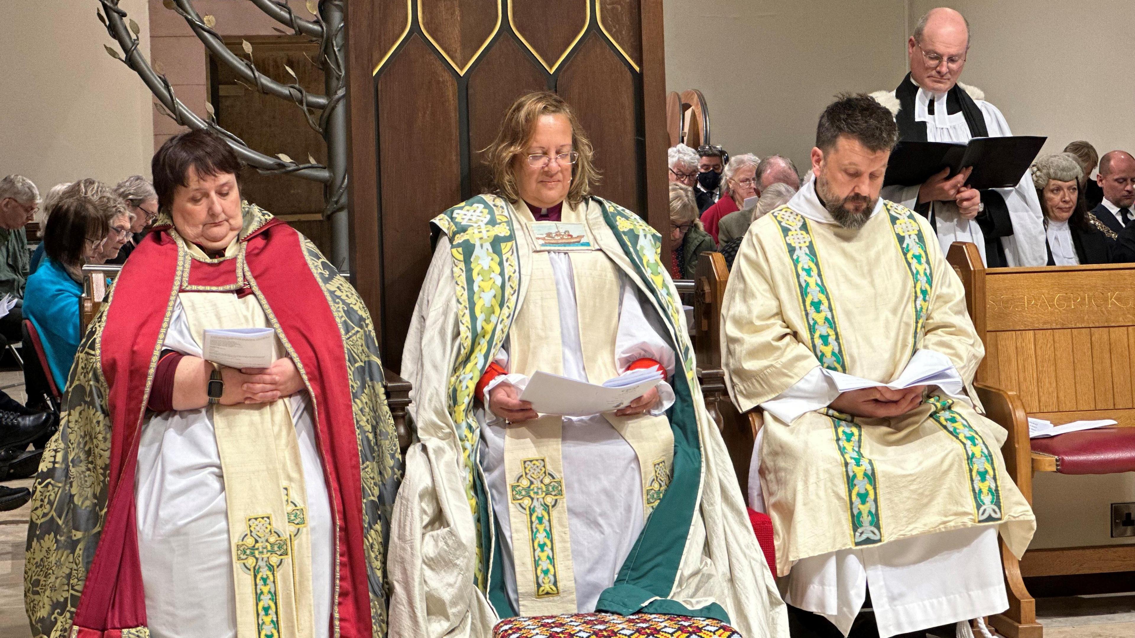 Right Reverend Patricia Hills siting in her throne in a beige, green and yellow vestment. There are other members of the clergy sitting either side, with the Archdeacon to the left in a gold and red vestment.