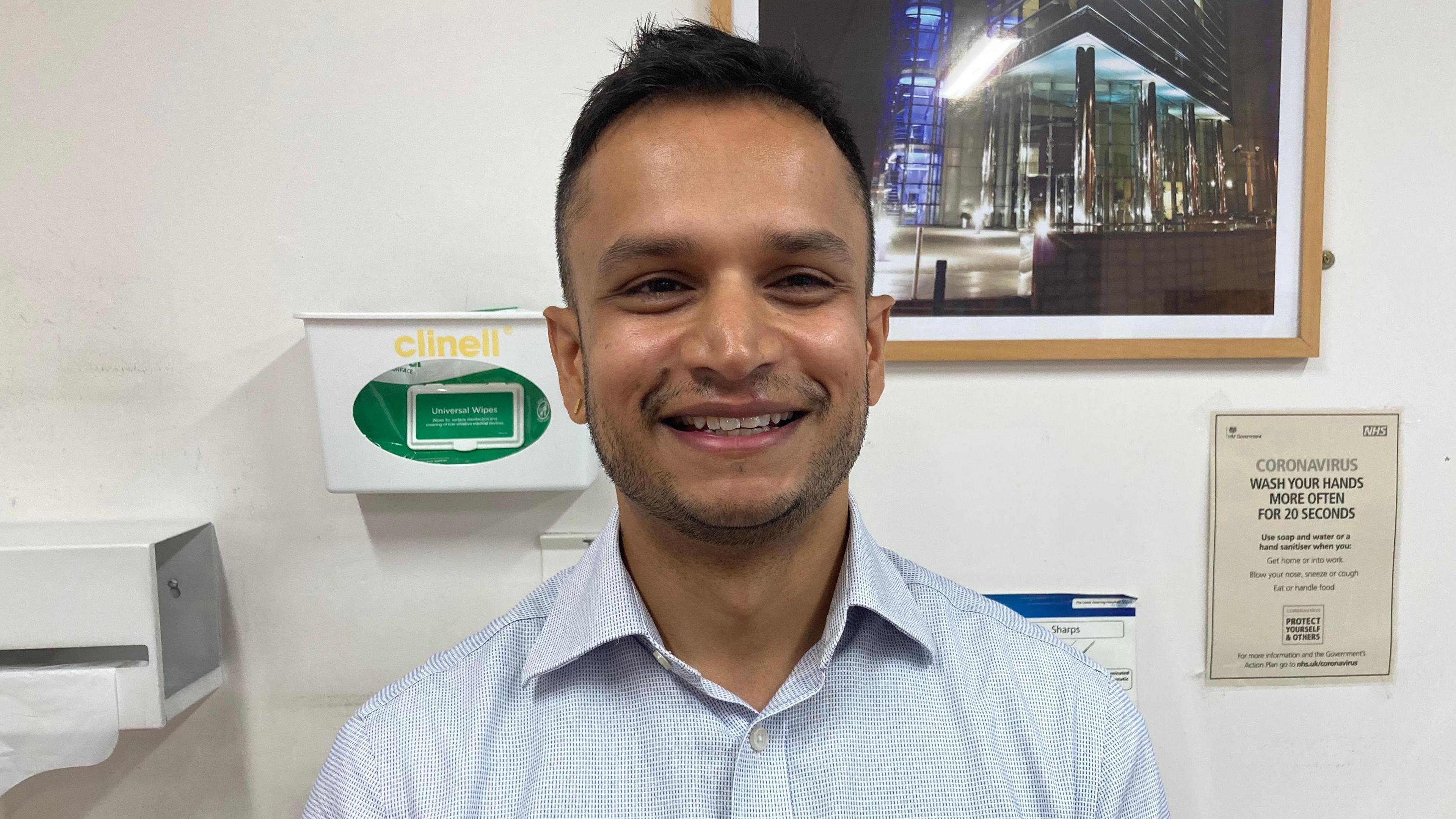 A man in a blue shirt smiles. He has dark hair and stubble. Behind him, a photo of a city scene hangs in a frame on the wall above a coronavirus poster.