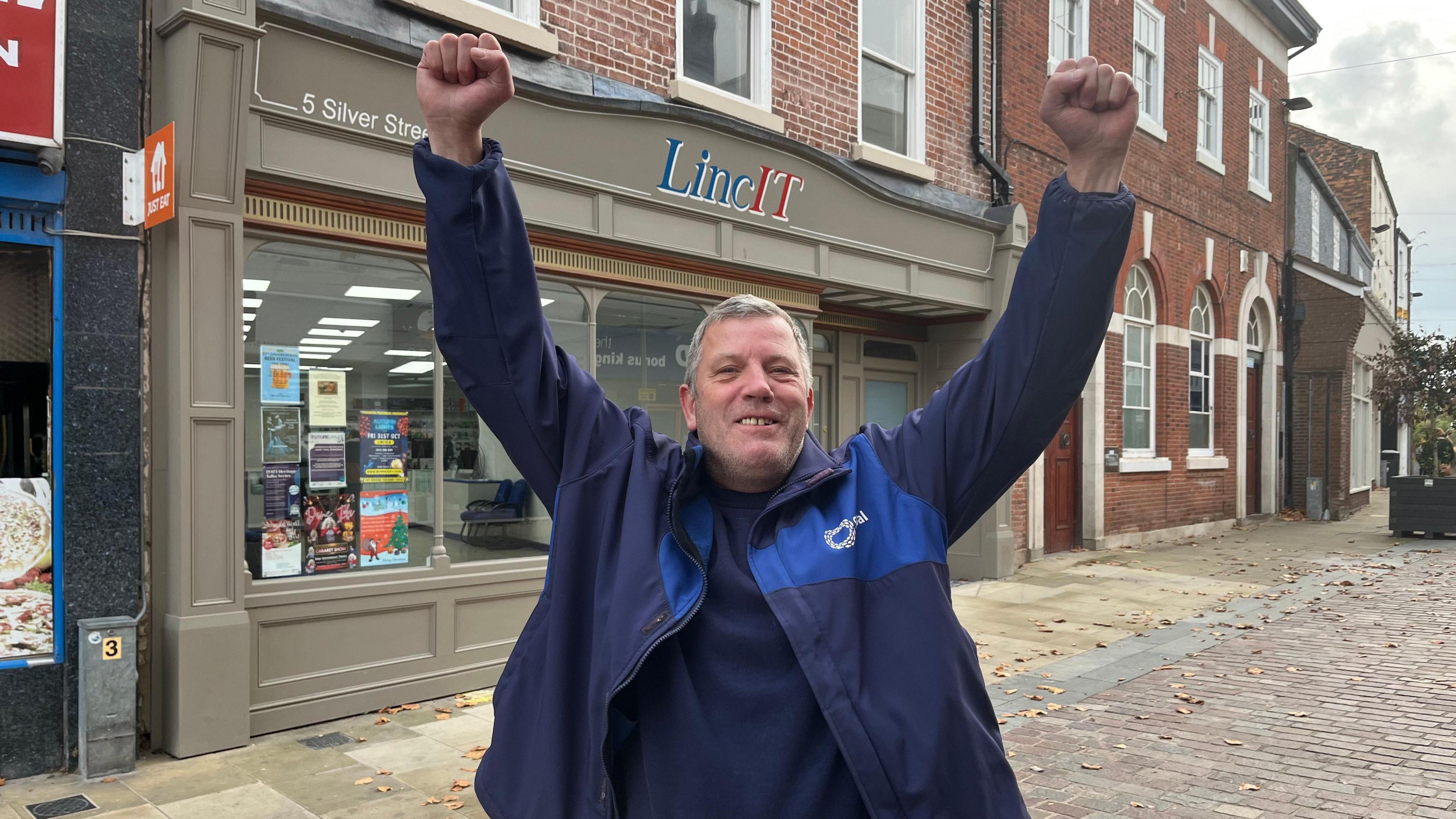 A man with short grey hair smiling while he puts both his hands in the air with his fists clenched. He is wearing a blue jacket and a blue top. Behind him is a row of buildings including one that has "LincIT" on the its sign.