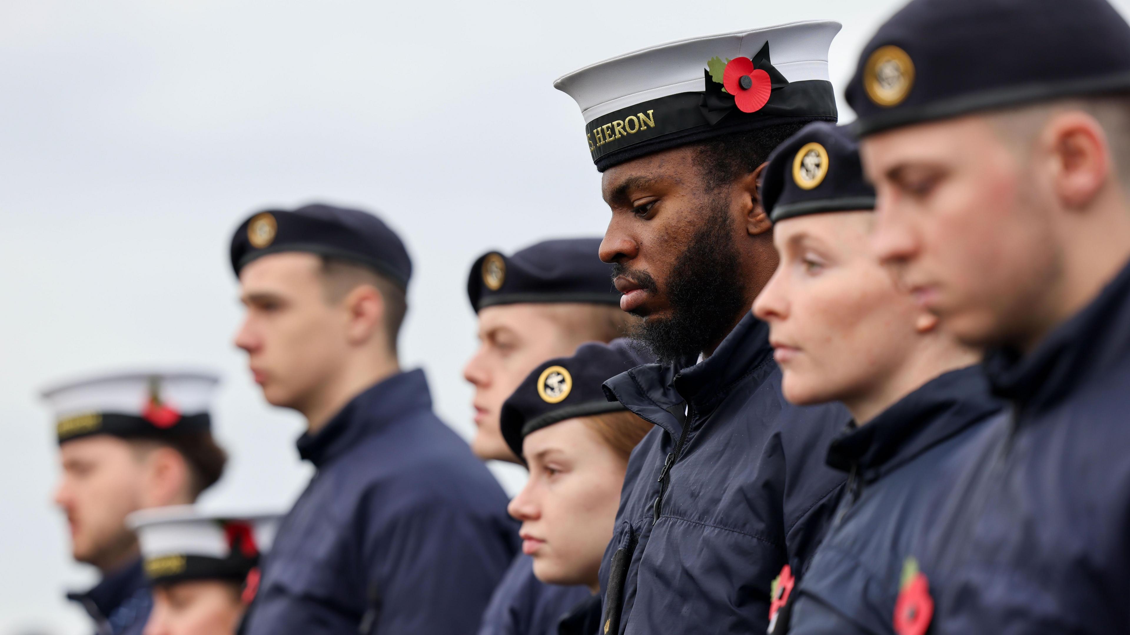 Navy soldiers in blue uniforms, both men and women, stand to attention in a line wearing poppies and uniform