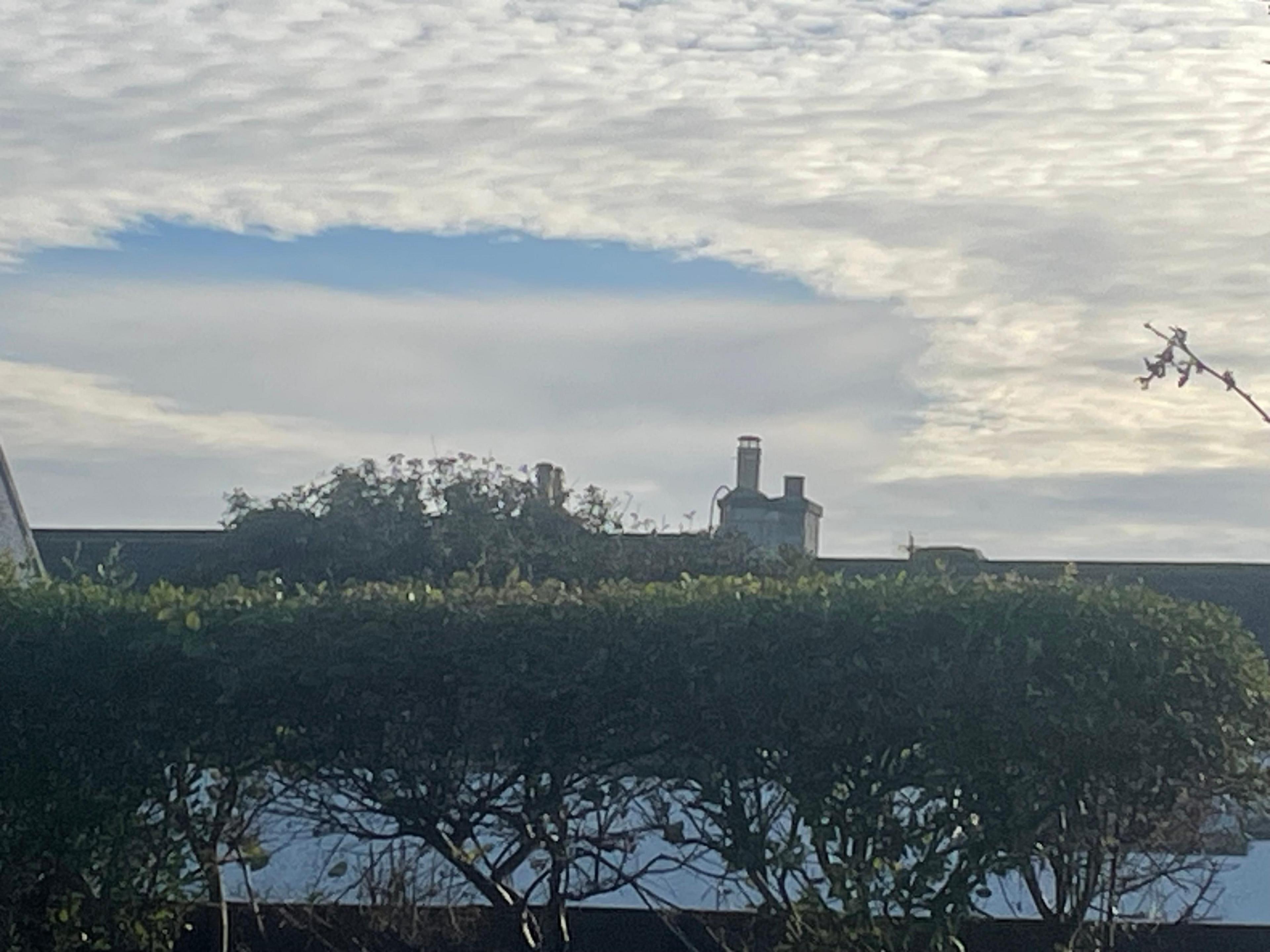 A large circular gap has formed in a layer of thin white cloud over the rooftops of Kingston, Moray. The blue sky is visible through the hole, with the edge of the cloud appearing sharply defined. In the foreground are neatly trimmed hedges and the top of a house chimney silhouetted against the sky.