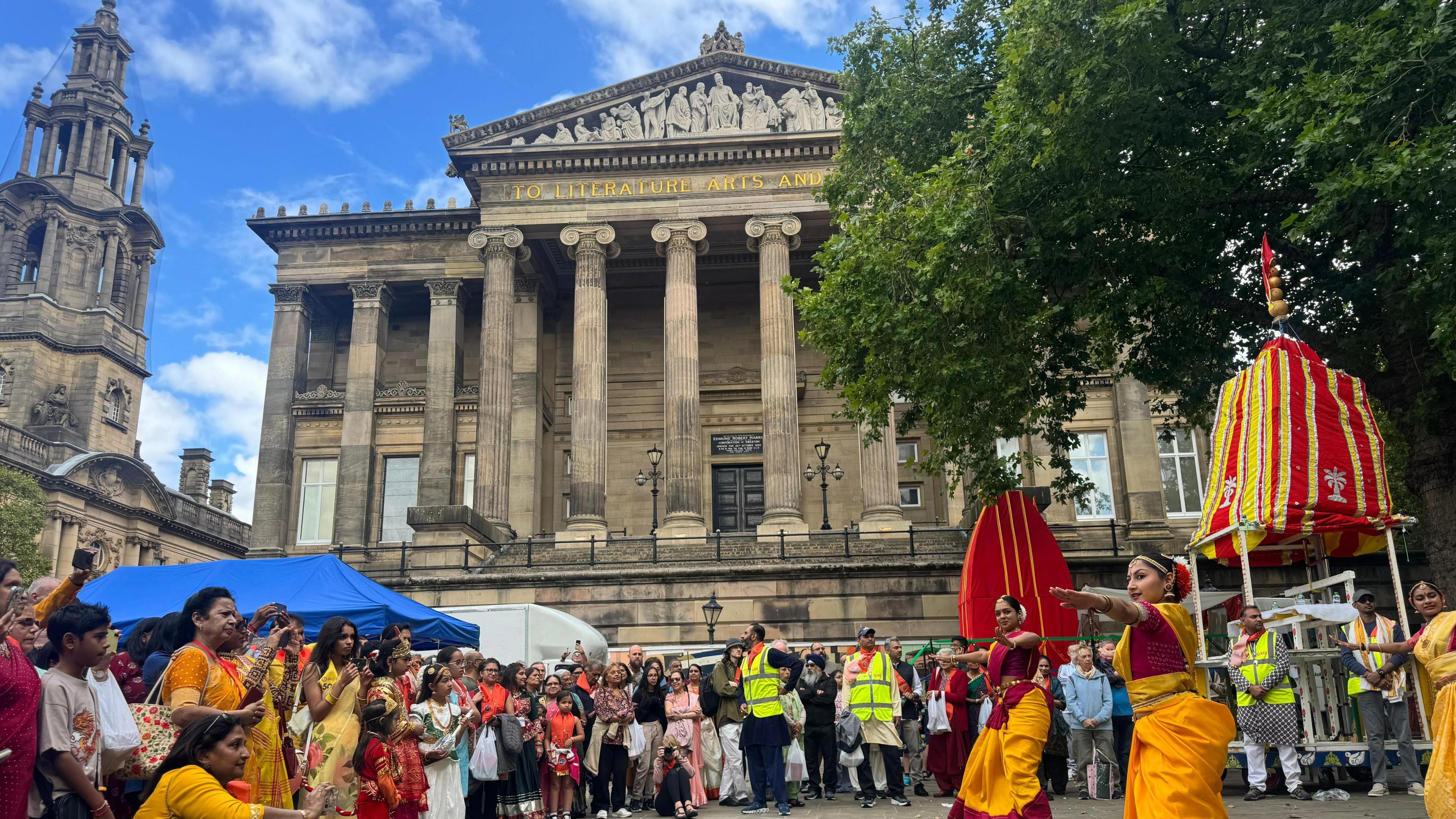 The classical pillared front of Harris Hall rises in the backdrop above crowds on the left watching some women on the right as they perform a traditional dance in red and yellow saris below trees.