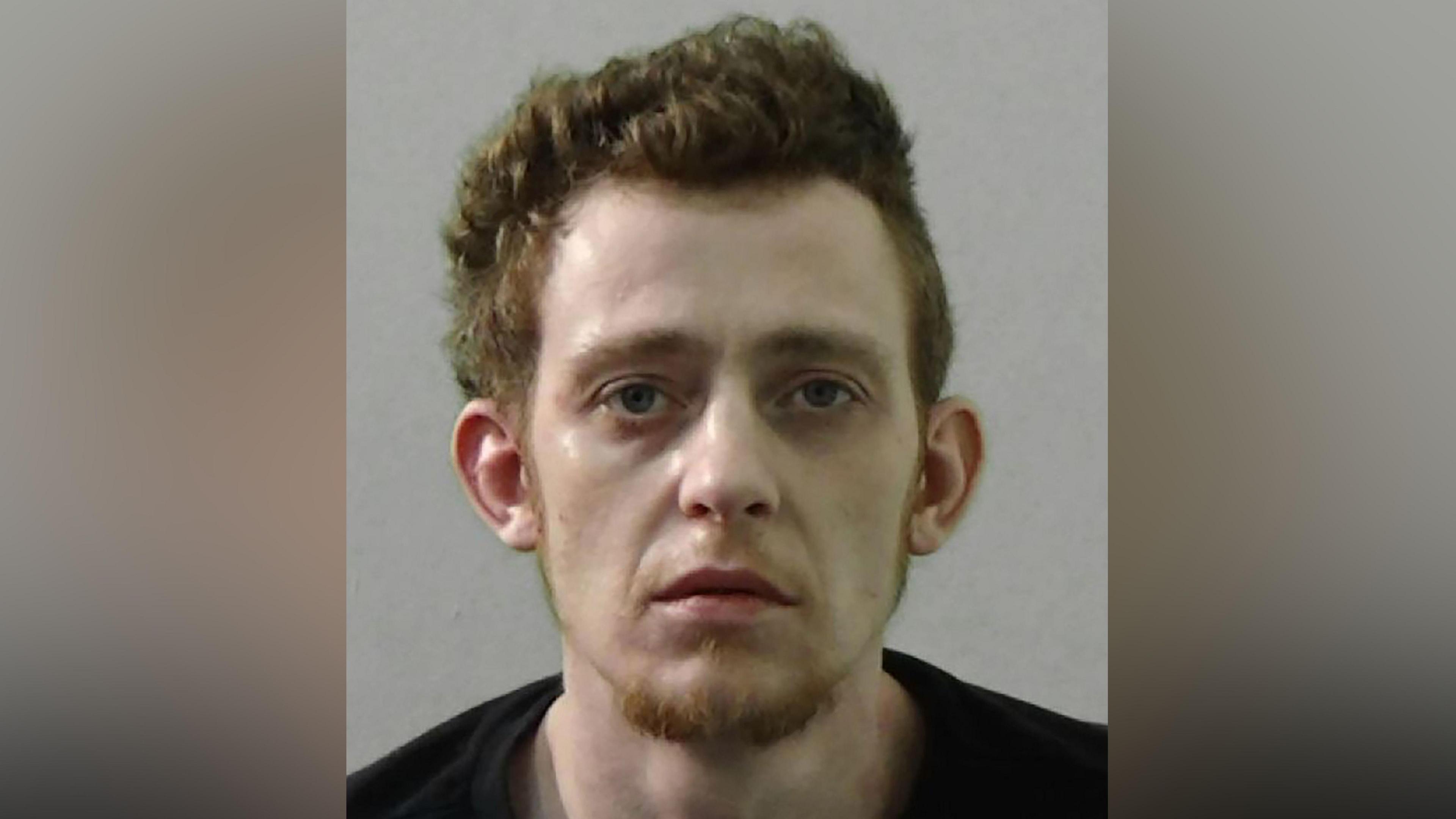 Mugshot of Reece Galbraith. He is gaunt and pale with thick ginger hair and a ginger goatee, wearing a black t shirt