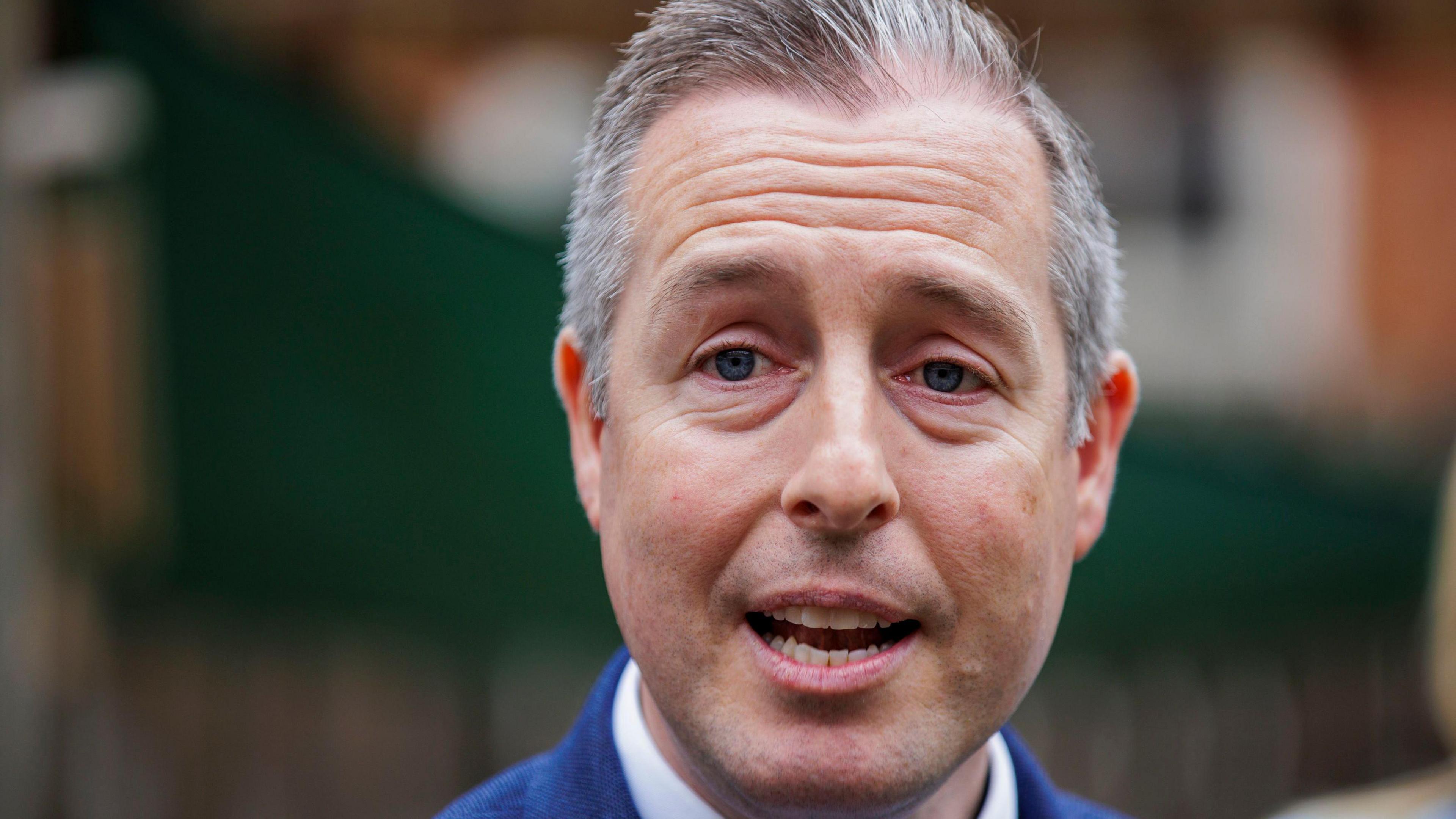 Paul Givan has short grey hair and is speaking in the image. He is wearing a navy suit and a white shirt. The background behind him is blurred.