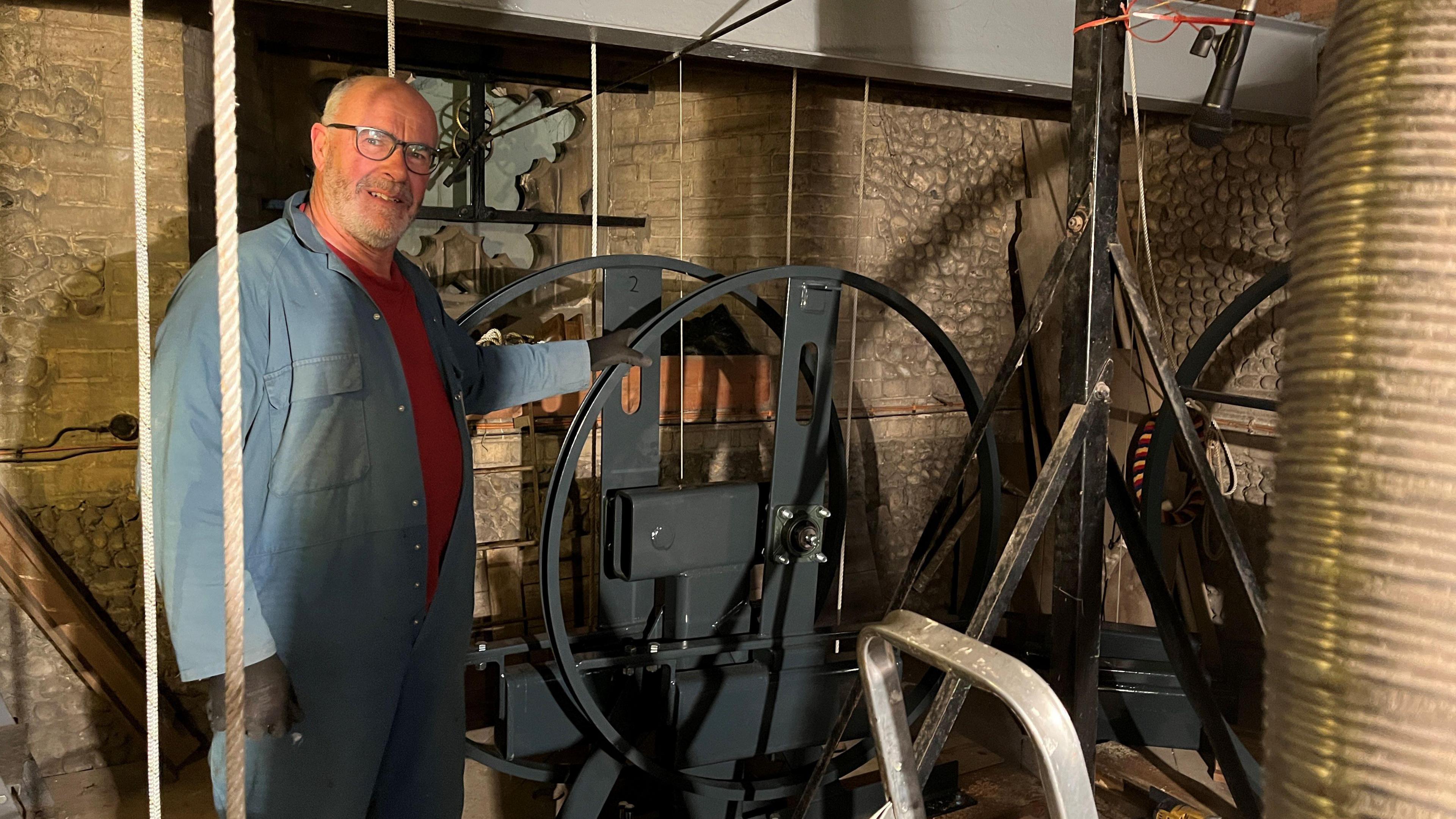 Neil Thomas stands inside a bell tower next to a large grey metal flywheel. He has very short grey hair and glasses, and is wearing blue overalls over a red top.