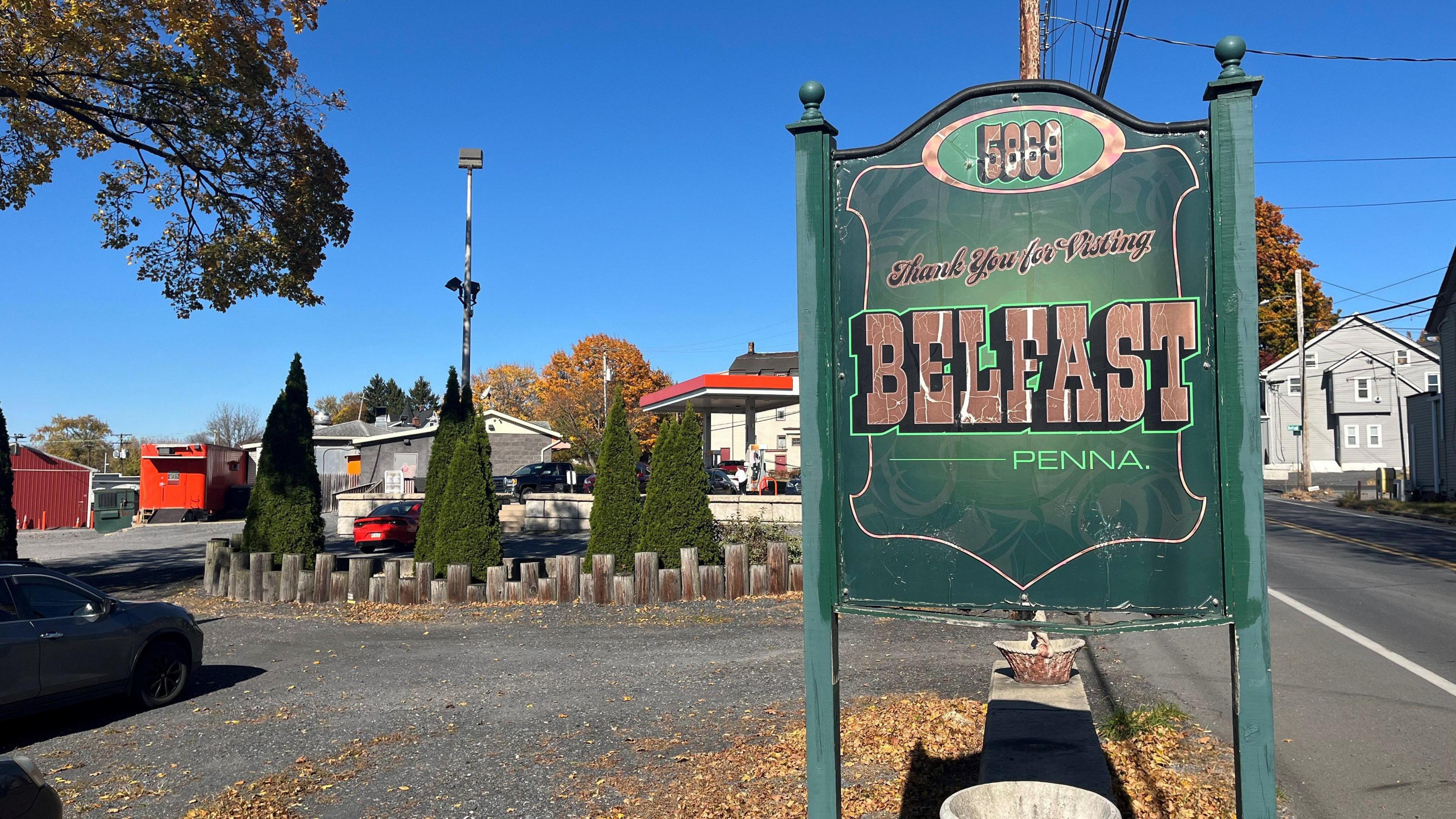 A sign saying thank you for visiting Belfast, Penna. It's green with brown writing. A grey wood house and some trees are in the background. Autumn leaves are lying on the road. The sky is clear and blue.