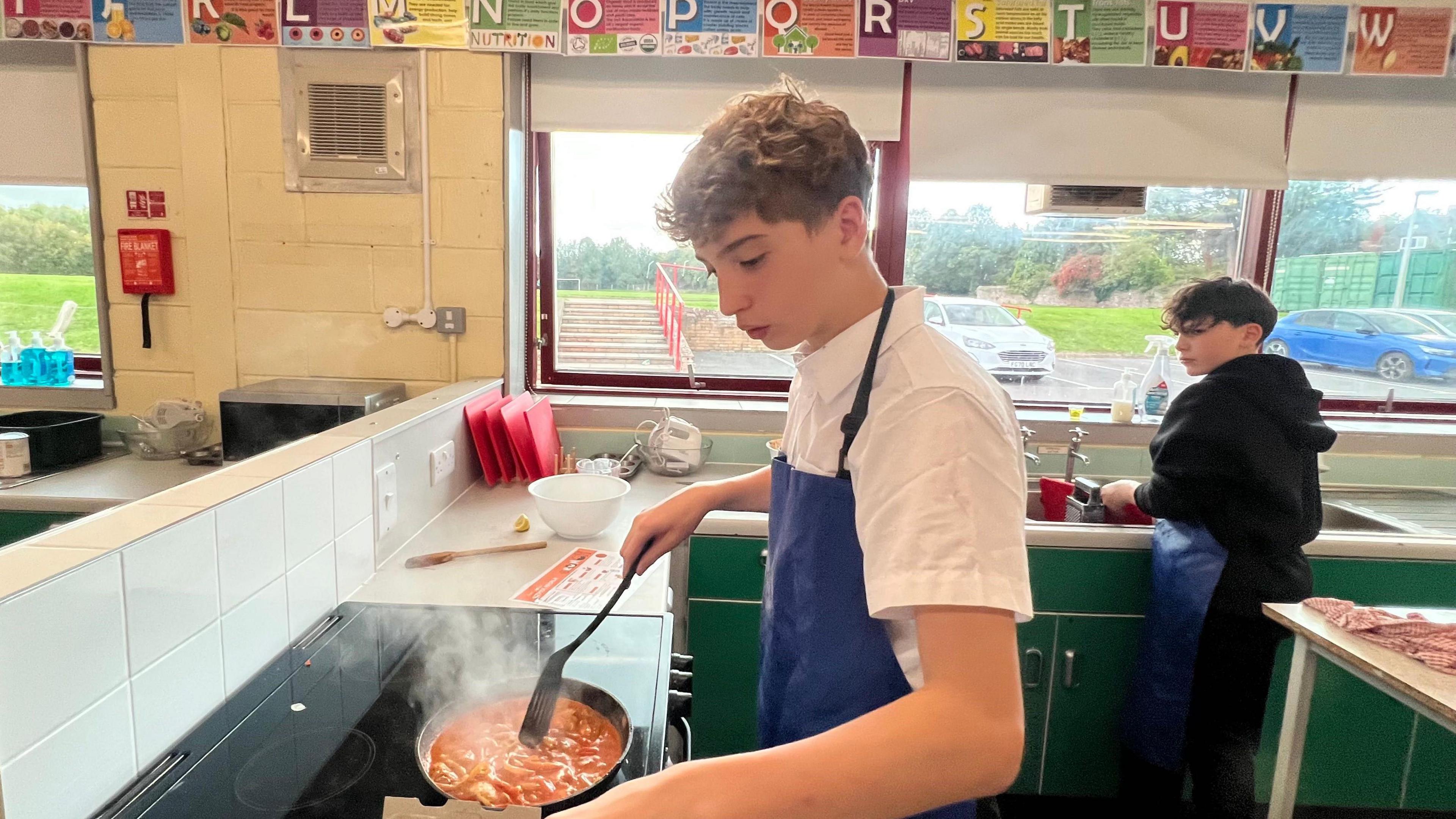 A school boy cooking chicken tikka masala at a stove. He is stirring a pot and is wearing a blue apron. He is looking down at the food, which is steaming hot. Another boy is in the background washing dishes.