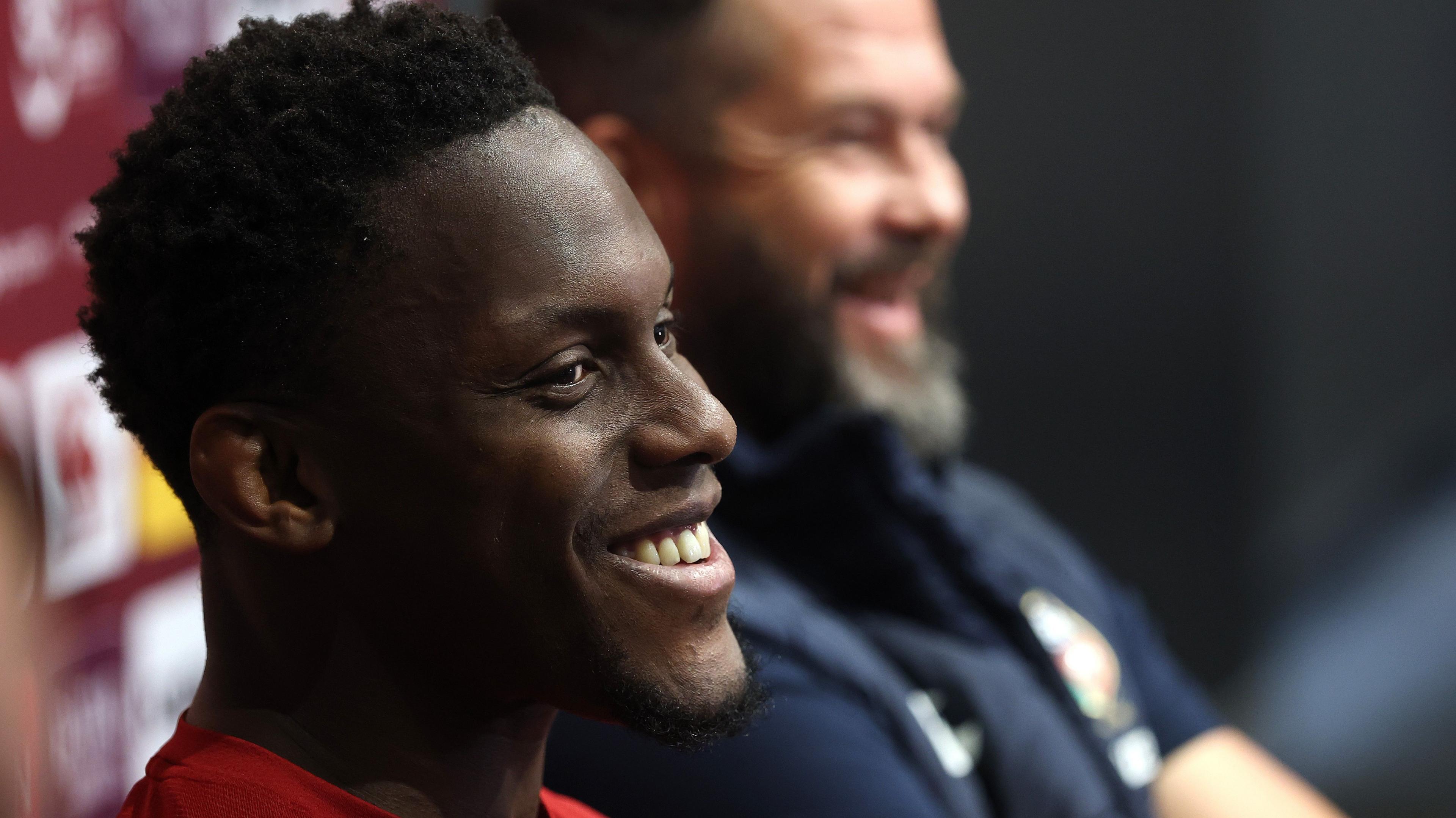 Maro Itoje, pictured sideways on, loos to the right of frame and smiles, with Andy Farrell, out of focus at the back of the shot, looking in the same direction and also smiling