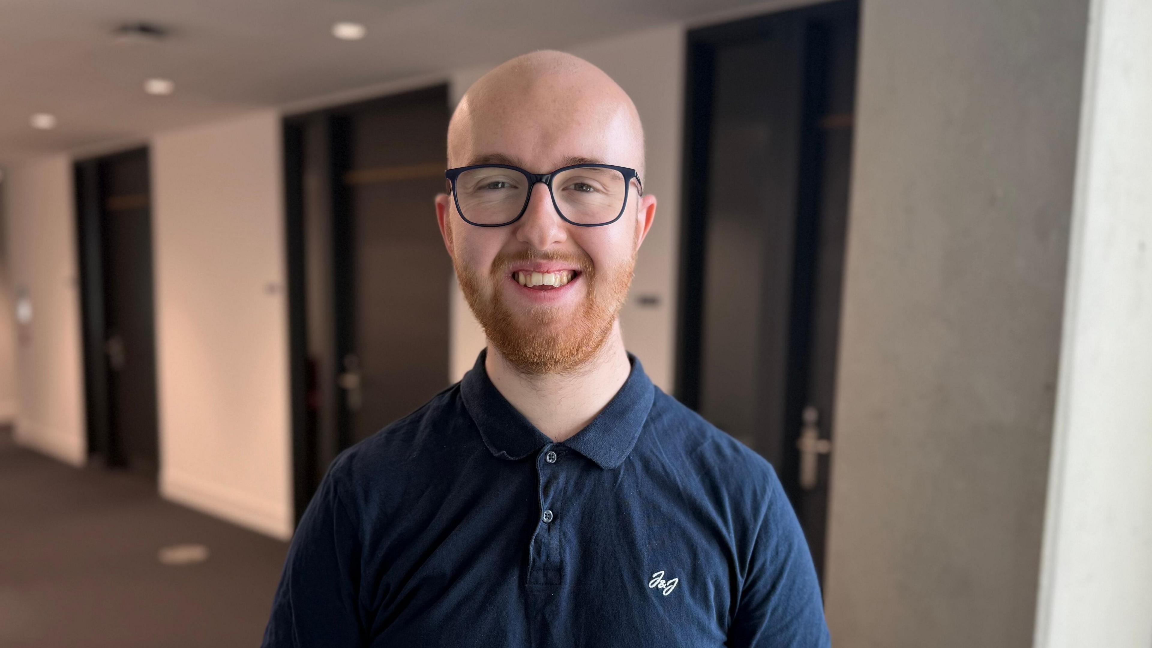 A young man standing in a hallway. He is wearing a blue polo shirt, and reading glasses.