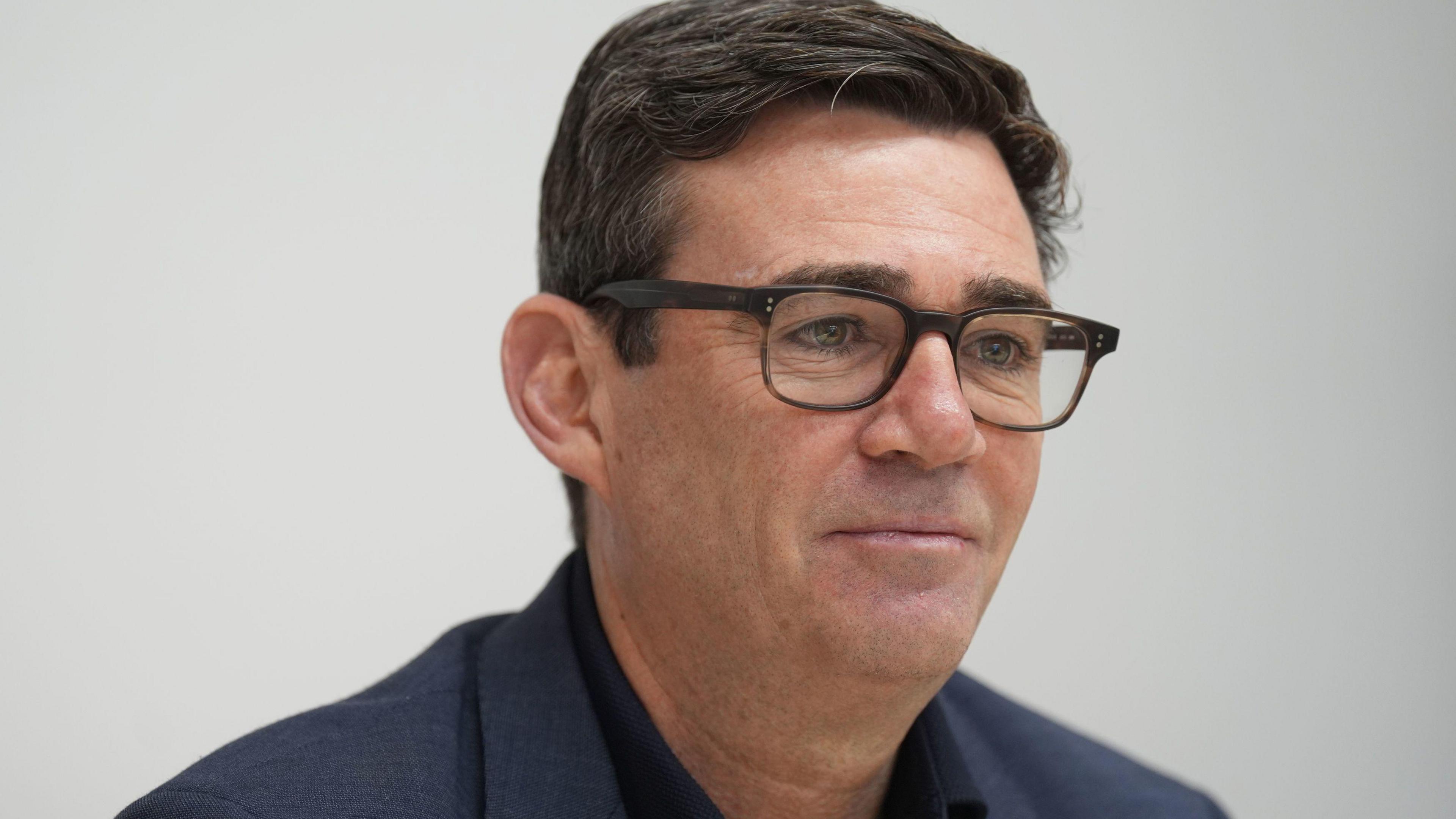 Clean shaven Andy Burnham with flecks of grey in his black hair, dark spectacles, a black shirt and RAF blue coloured jacket.