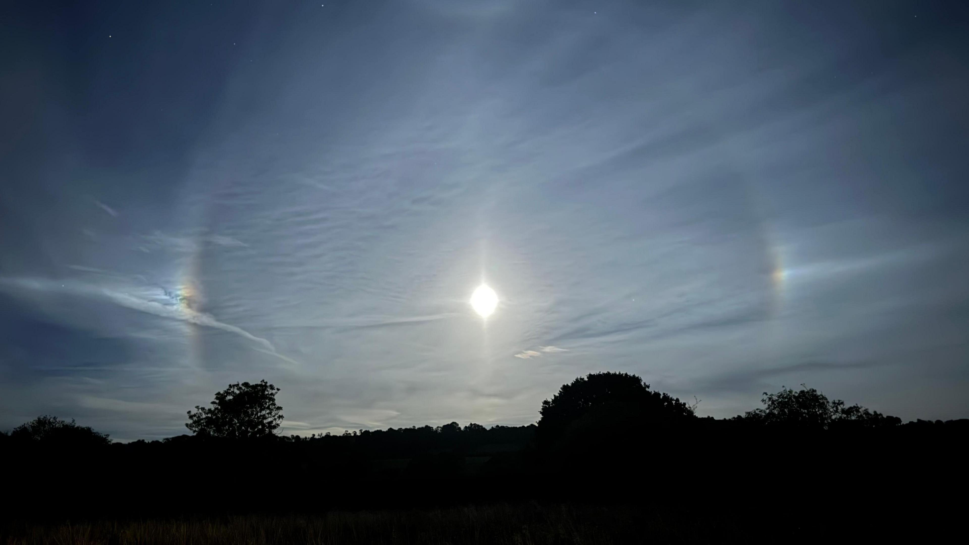 A moon in the night sky on a clear night surrounded by a rainbow orb. A line of trees can be seen beneath it.