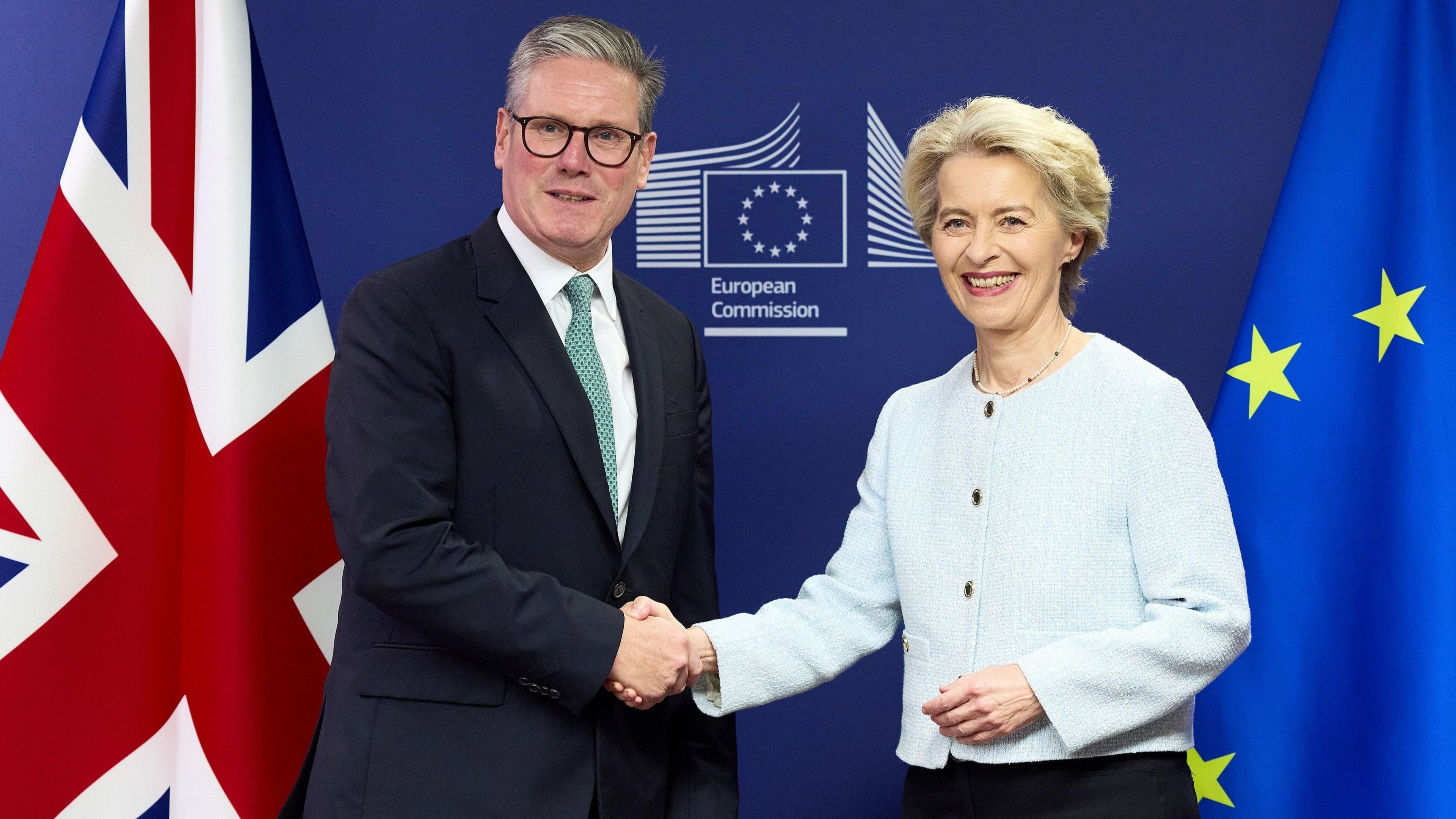 Prime Minister Keir Starmer in a black suit shakes hands with European Commission president Ursula von der Leyen in a pale blue blazer in front of a sign that says "European Commission" and the UK and EU flags at the European Commission headquarters in Brussels, Belgium