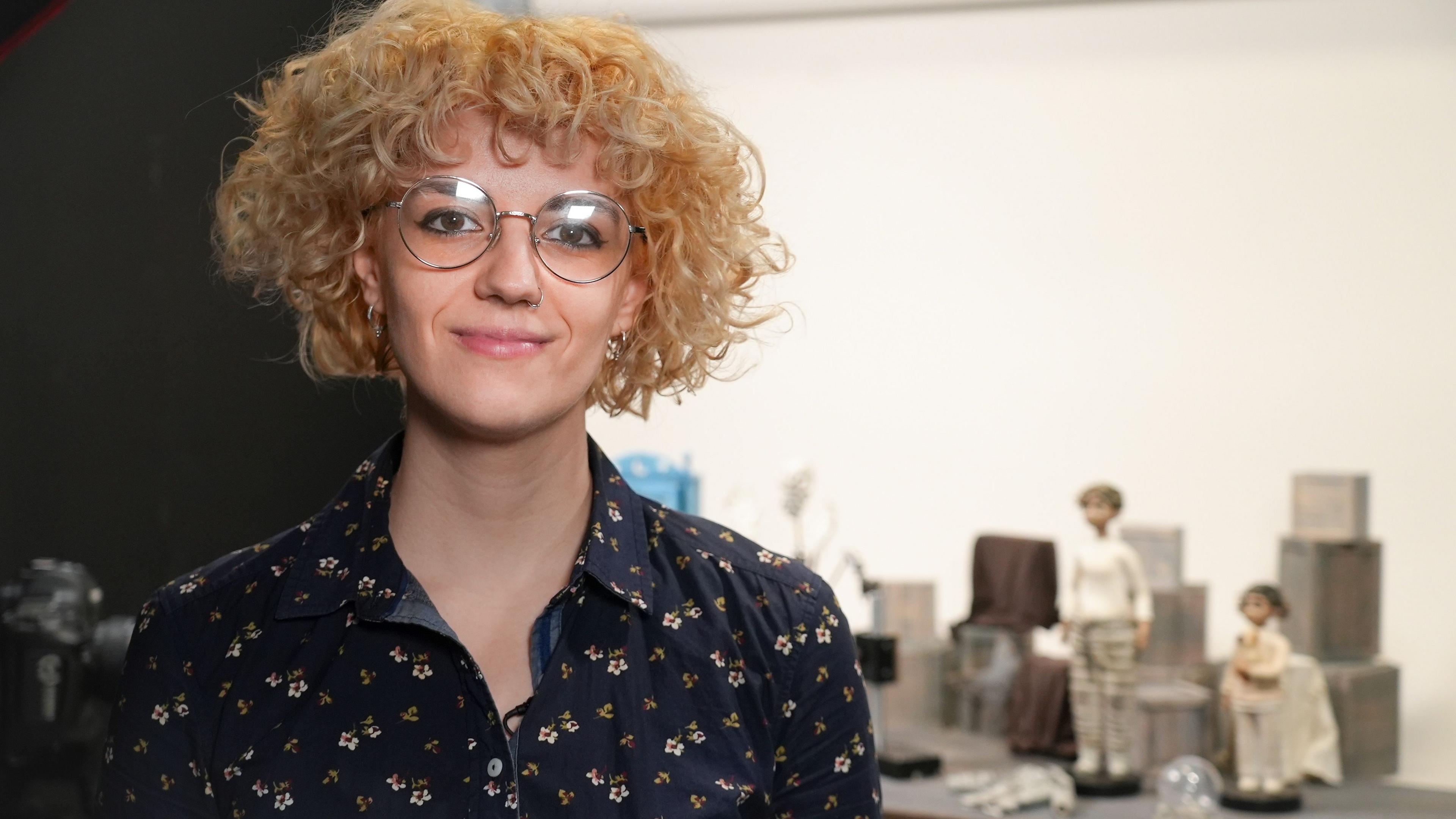 Laura Tofarides. Mae ganddi wallt golau a chyrliog ac mae'n gwisgo sbectol a chrys blodeuog.