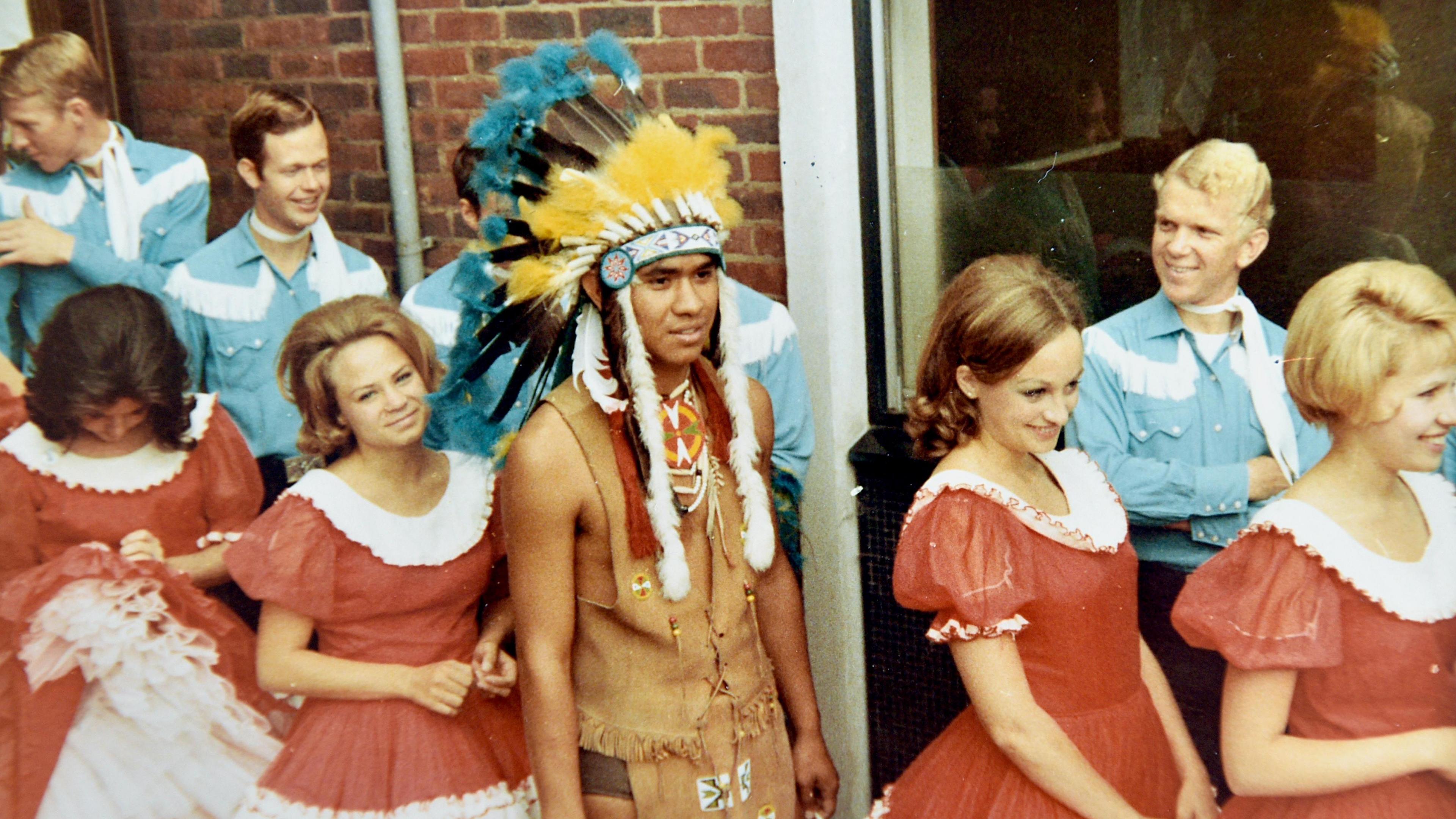 A 1960s picture showing several young women in red and white dresses with young men in background wearing light blue shirts and talking to each other. They are lined up alongside a brick building. A young man in middle of photo is wearing a Native American outfit with traditional headdress.