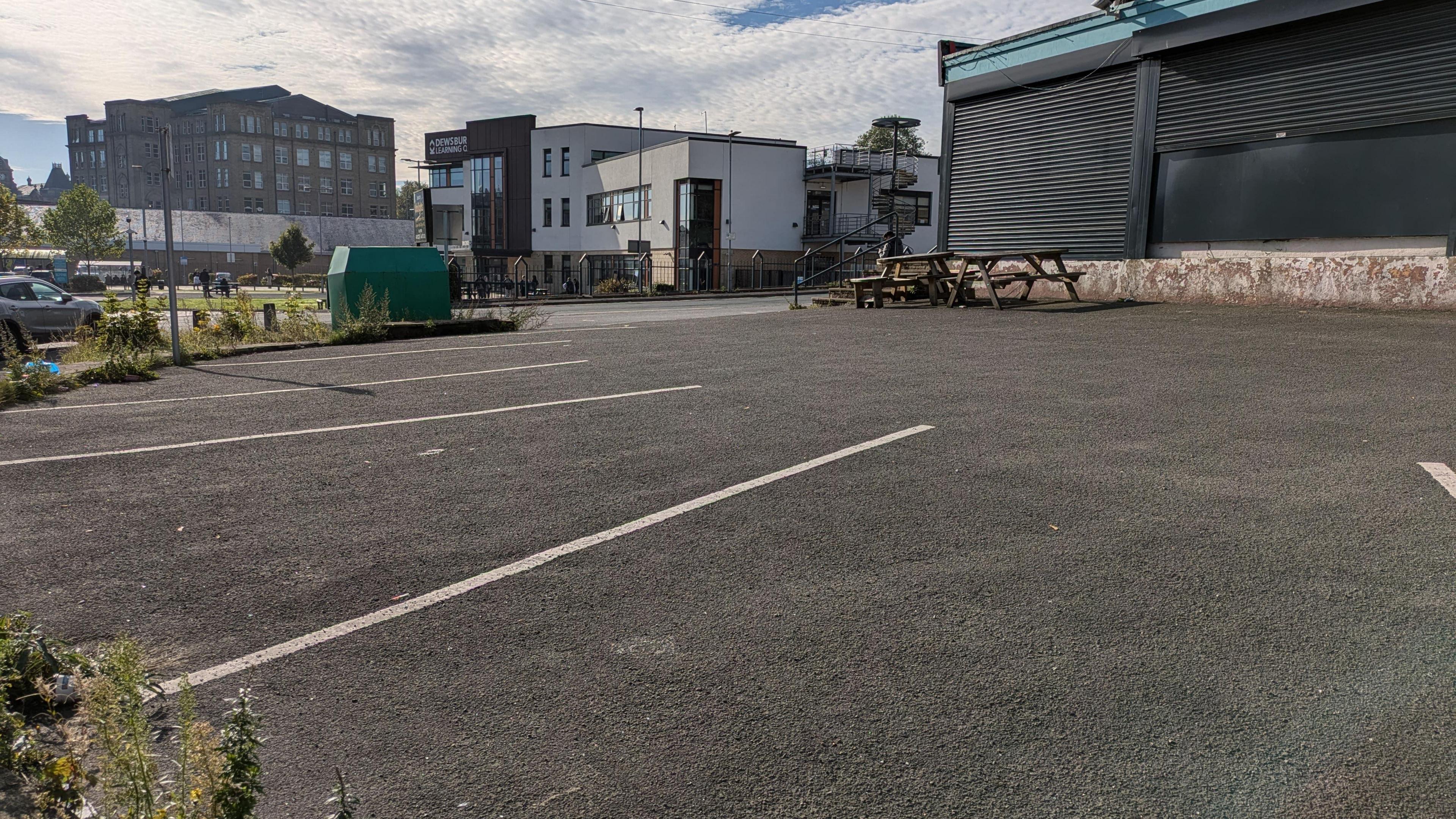 An empty car park on sunny day. Pictured are spaces without cars in them, while a closed premises with its shutters down stands in the background, with a picnic table just in front.