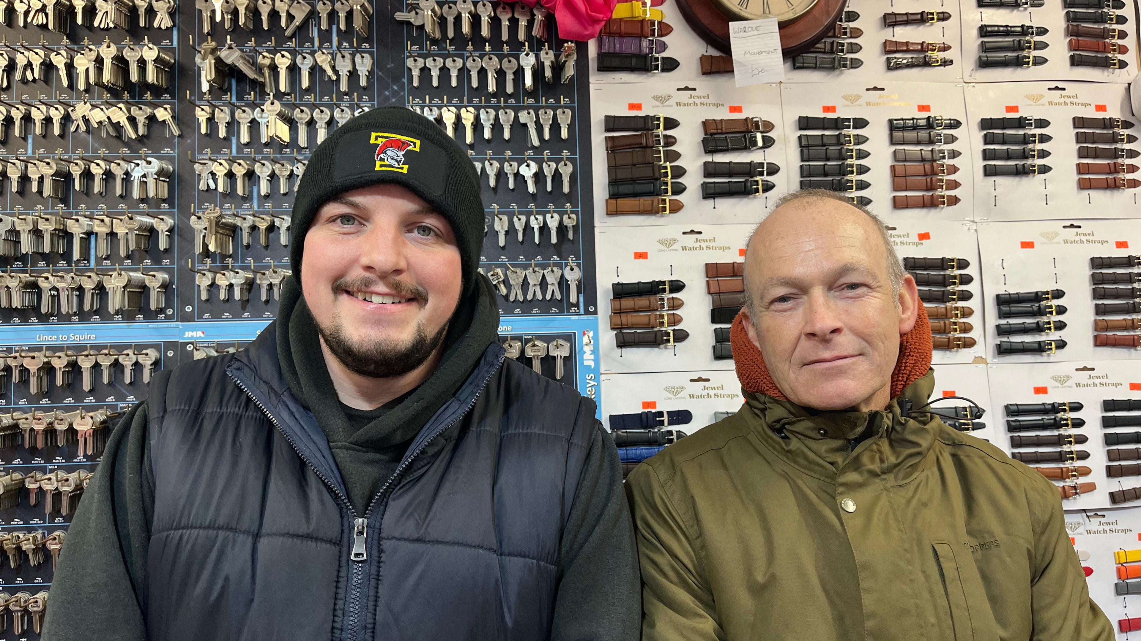 Aaron and Marcus side by side behind their stall, both looking into the camera. Aaron is wearing a navy puffa jacket, and Marcus is wearing a khaki raincoat. Behind them you can see rows and rows of keys ready to be cut, and a wall showing off the watch straps they sell. 