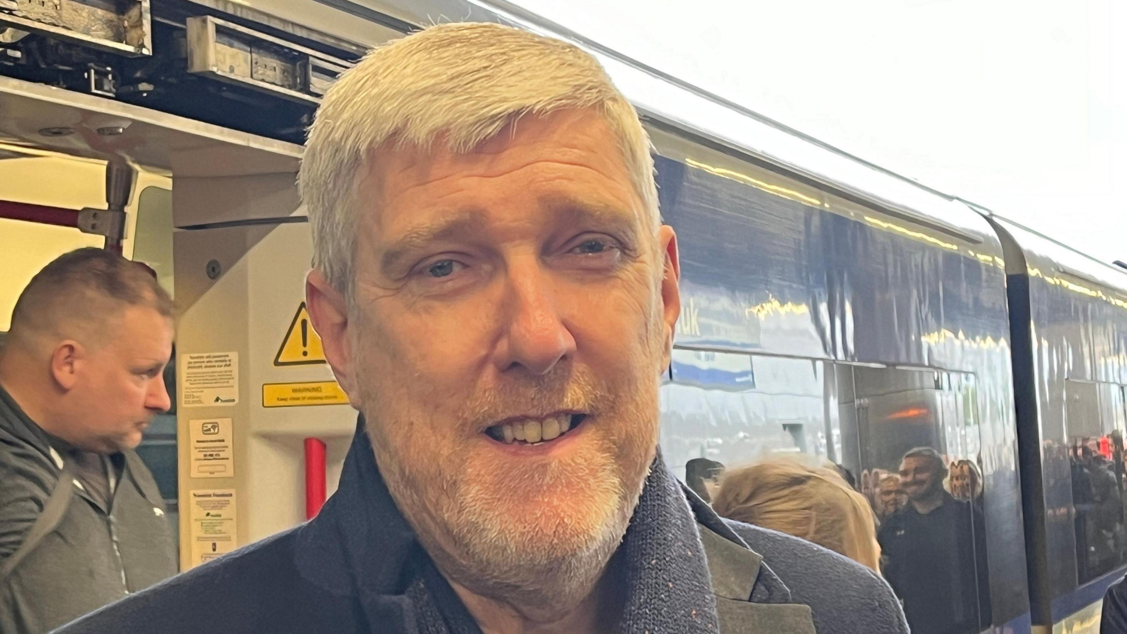 John O’Dowd is standing outside a train. He has white hair and is wearing a navy coat.