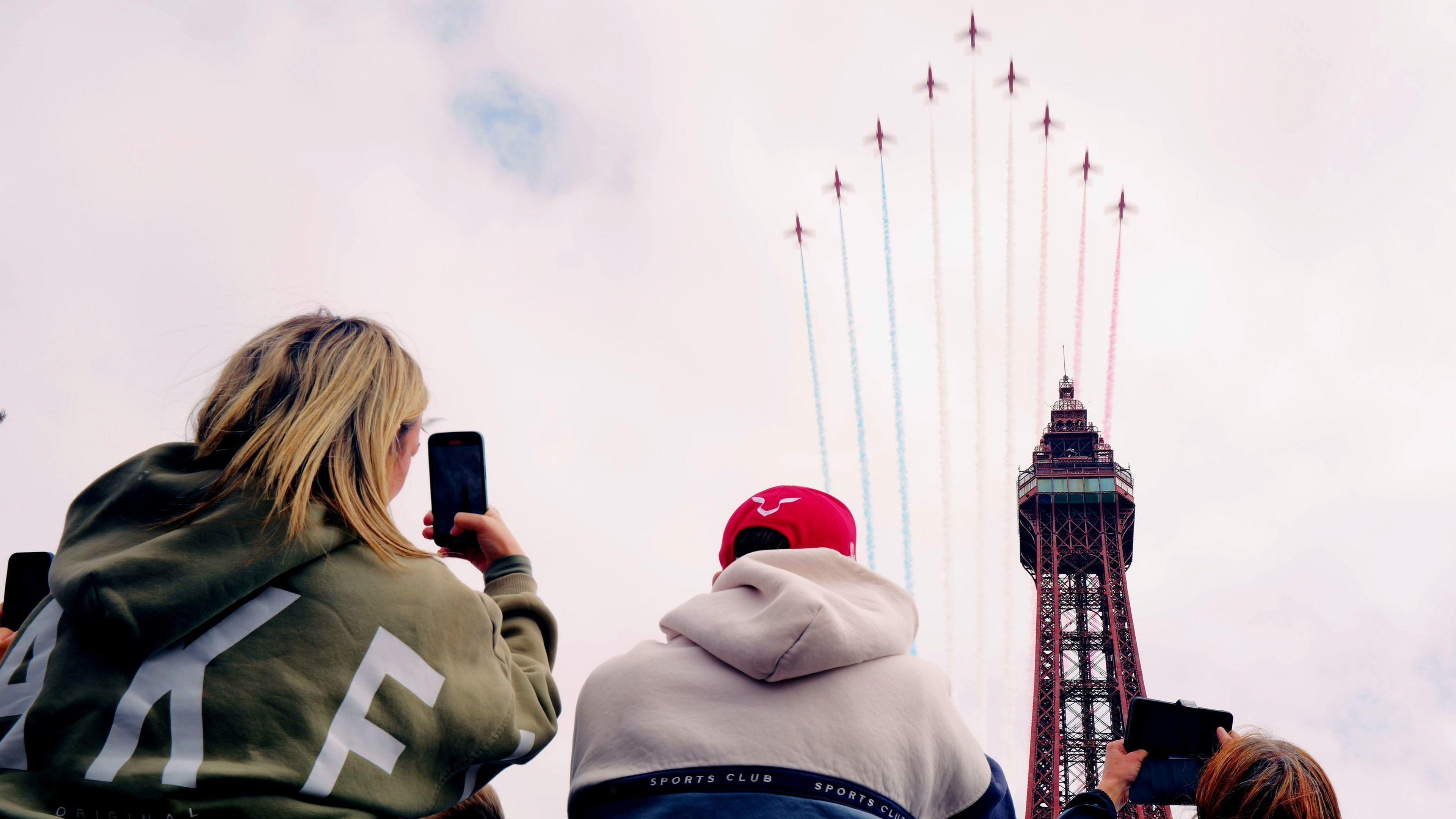 Ground view of an arrowhead formation of nine Red Arrows planes streaming blue, white and red plumes as they fly over Blackpool Tower while, at the bottom of the image, three people take pictures of it on their mobile phones.