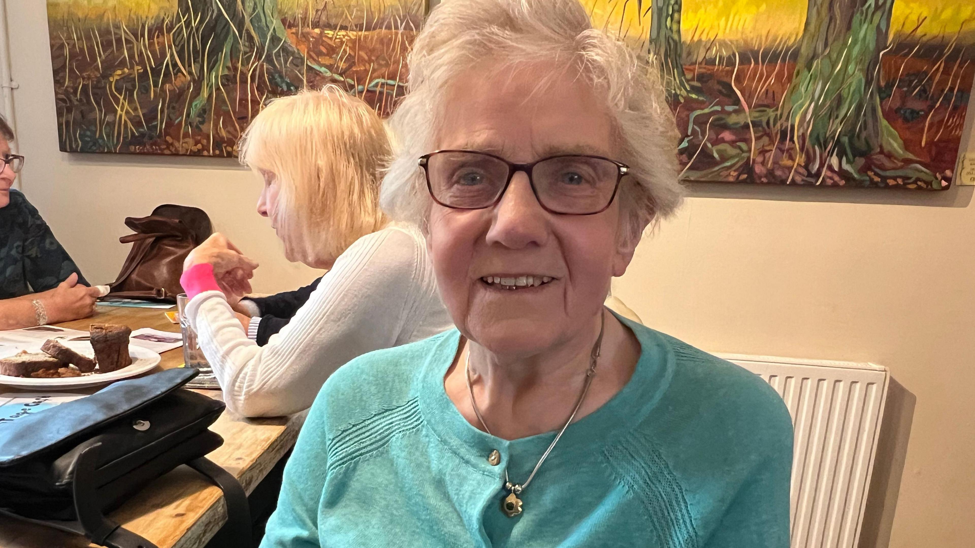 Marlene wearing glasses and a pale blue cardigan and is sitting next to a table with a cake on it and two other people talking across it