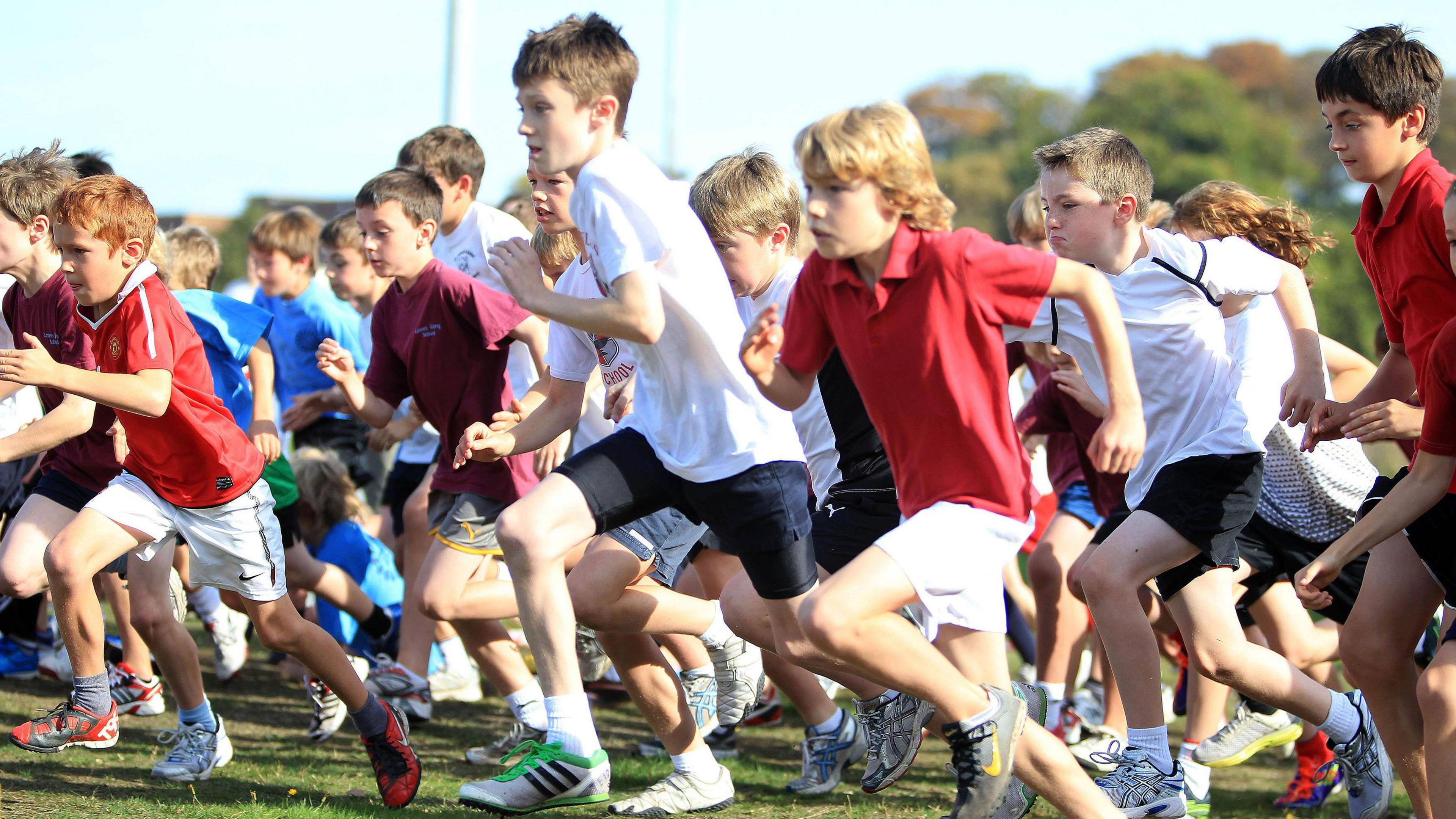 Photograph shows a large group of primary school children running together. Each of them are wearing sportswear and are running from right to left in a field.