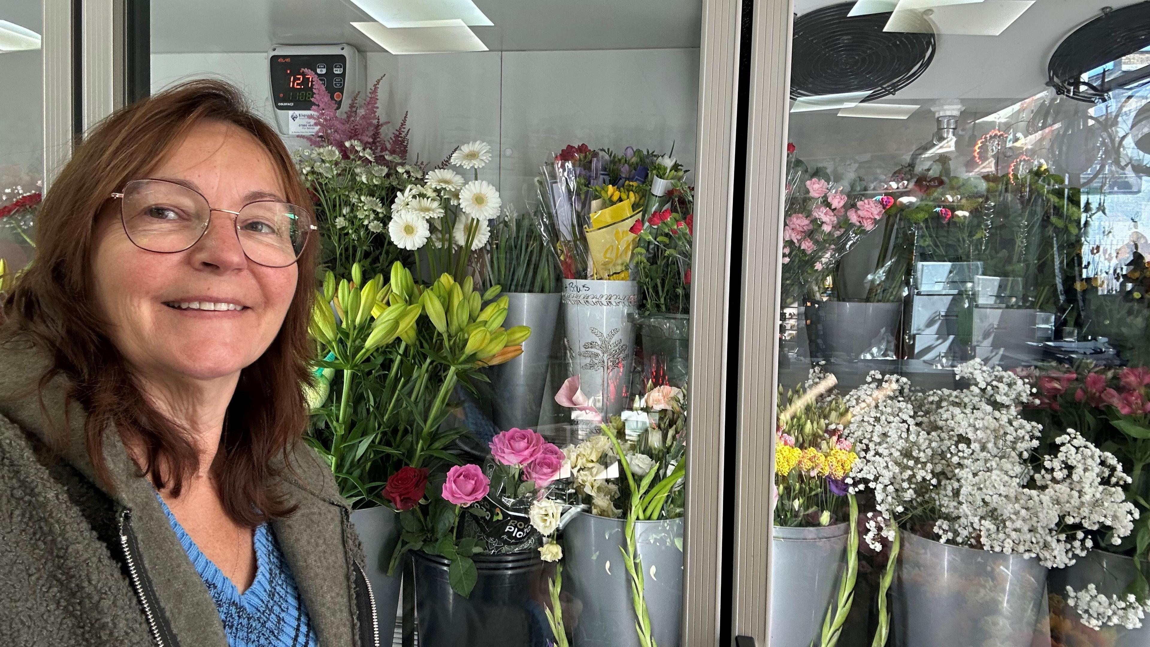 Bonnie Twigg, 58, has brown shoulder-length hair with a central parting. She is wearing brown-framed glasses and a blue and black 'v' necked sweater under a fleece. Behind her are an assortment of flowers in buckets and vases, on display in her shop.