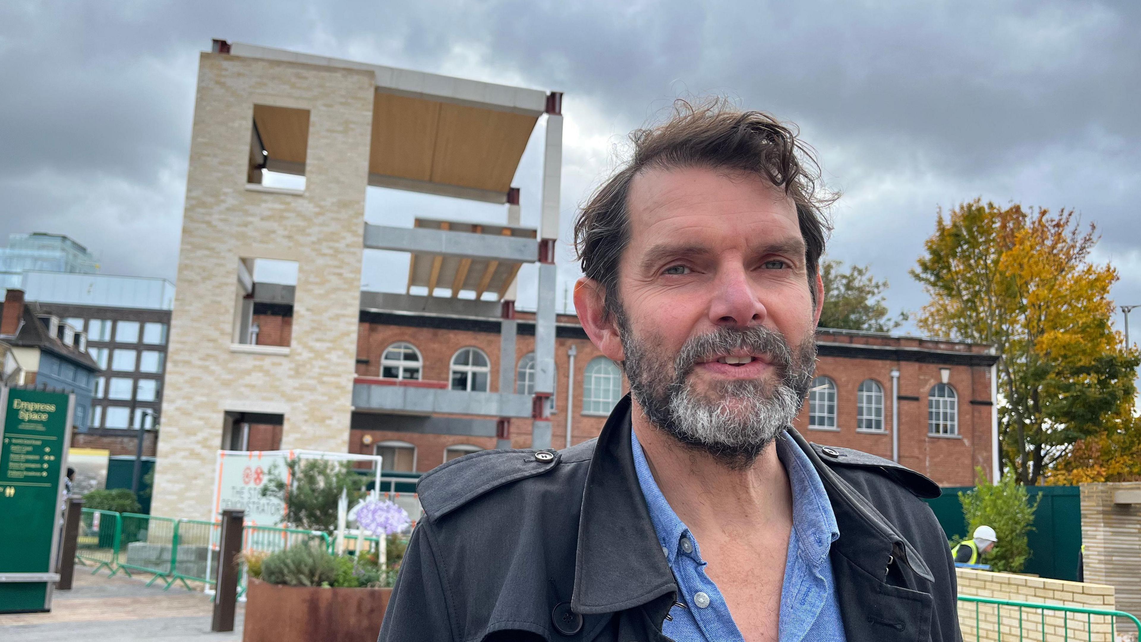 A man stands in front of a stone structure. He is wearing a blue shirt and looking into the camera.