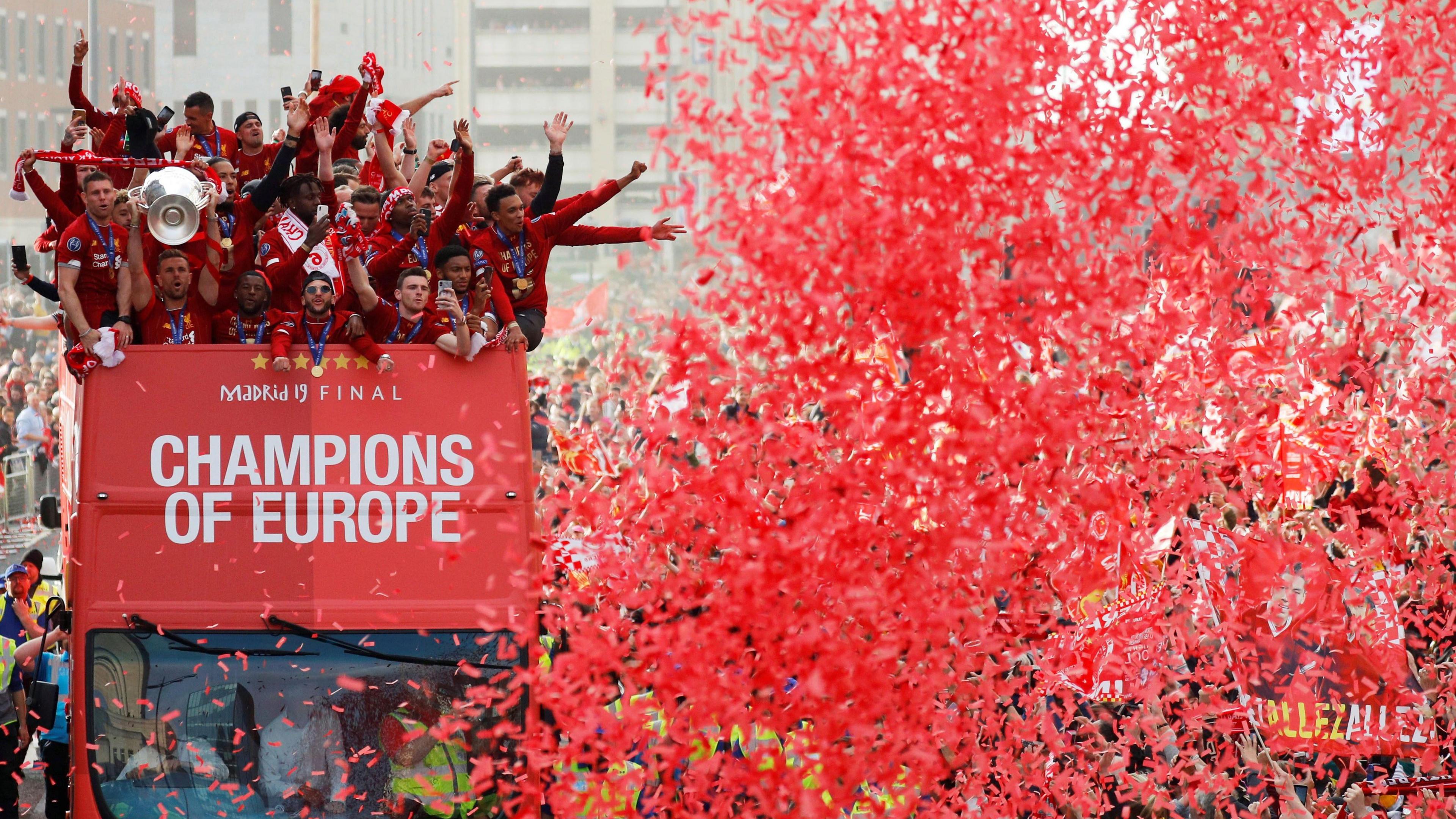 Red confetti in the air in front of the Liverpool bus in 2019. Players raise their arms in celebration on the open-top bus.