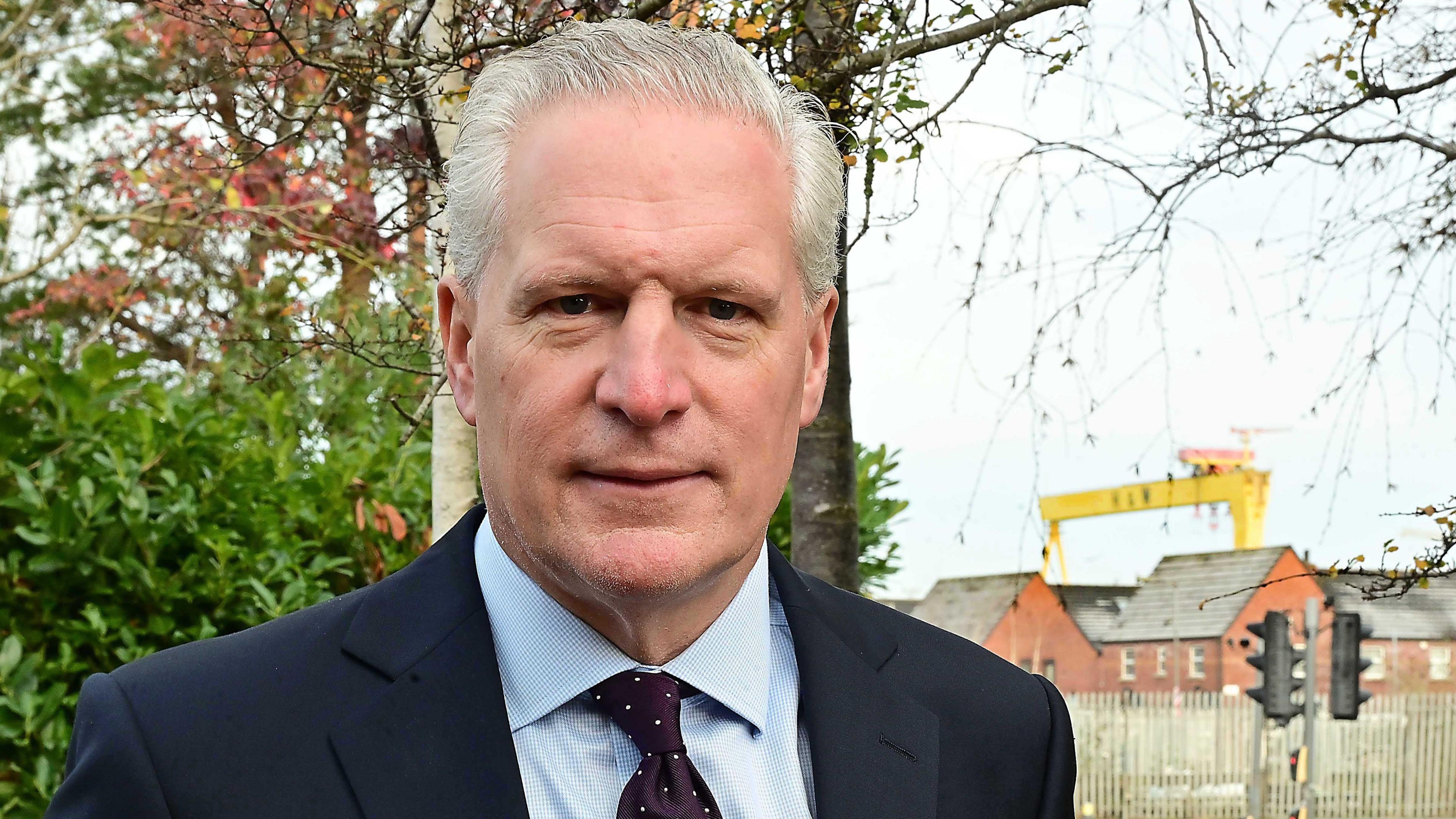 Alan Stout. He has short grey hair, wearing a navy blazer, light blue shirt and navy polka dot tie. The Harland and Wolf cranes are in the background.