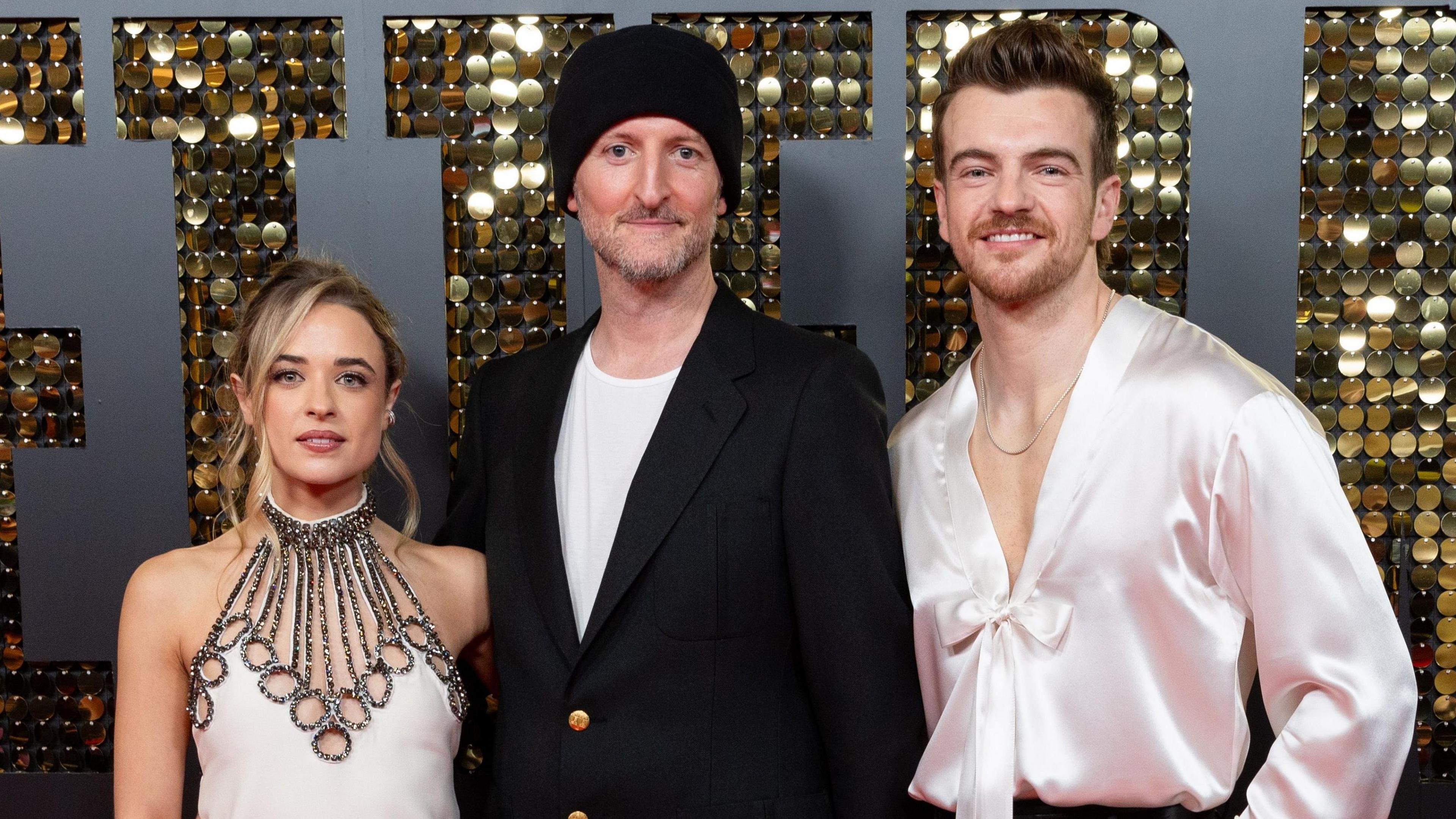 Raechelle Banno is dressed in a white dress, Michael Gracey has a black blazer over a t-shirt and Jonno has a silk shirt. They are at a premiere event for Better Man.