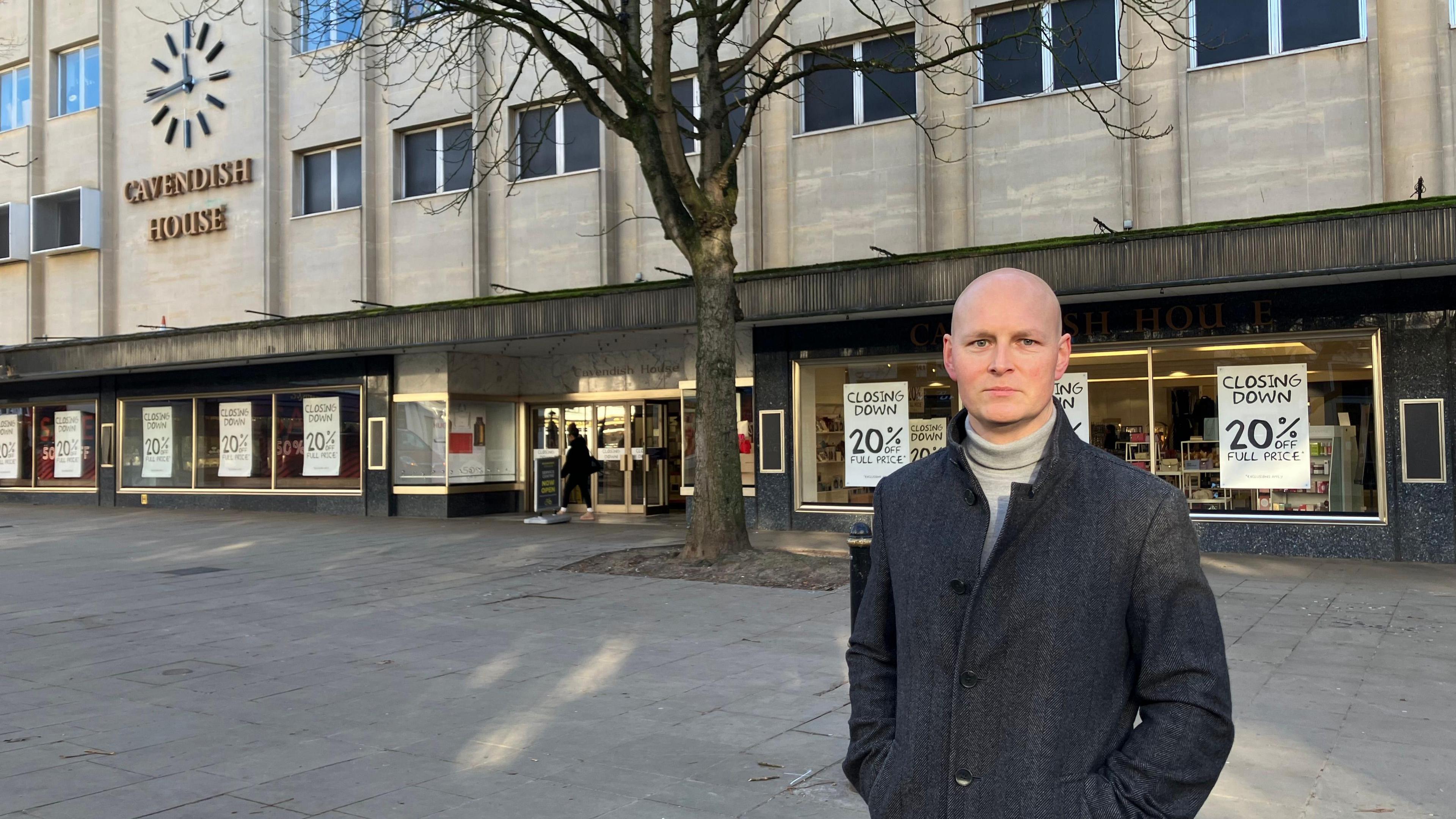 A bald middle aged man is wearing a long dark grey coat with a pale turtleneck beneath it as he looks sternly at the camera outside a department shop called Cavendish House on a winter's day.
