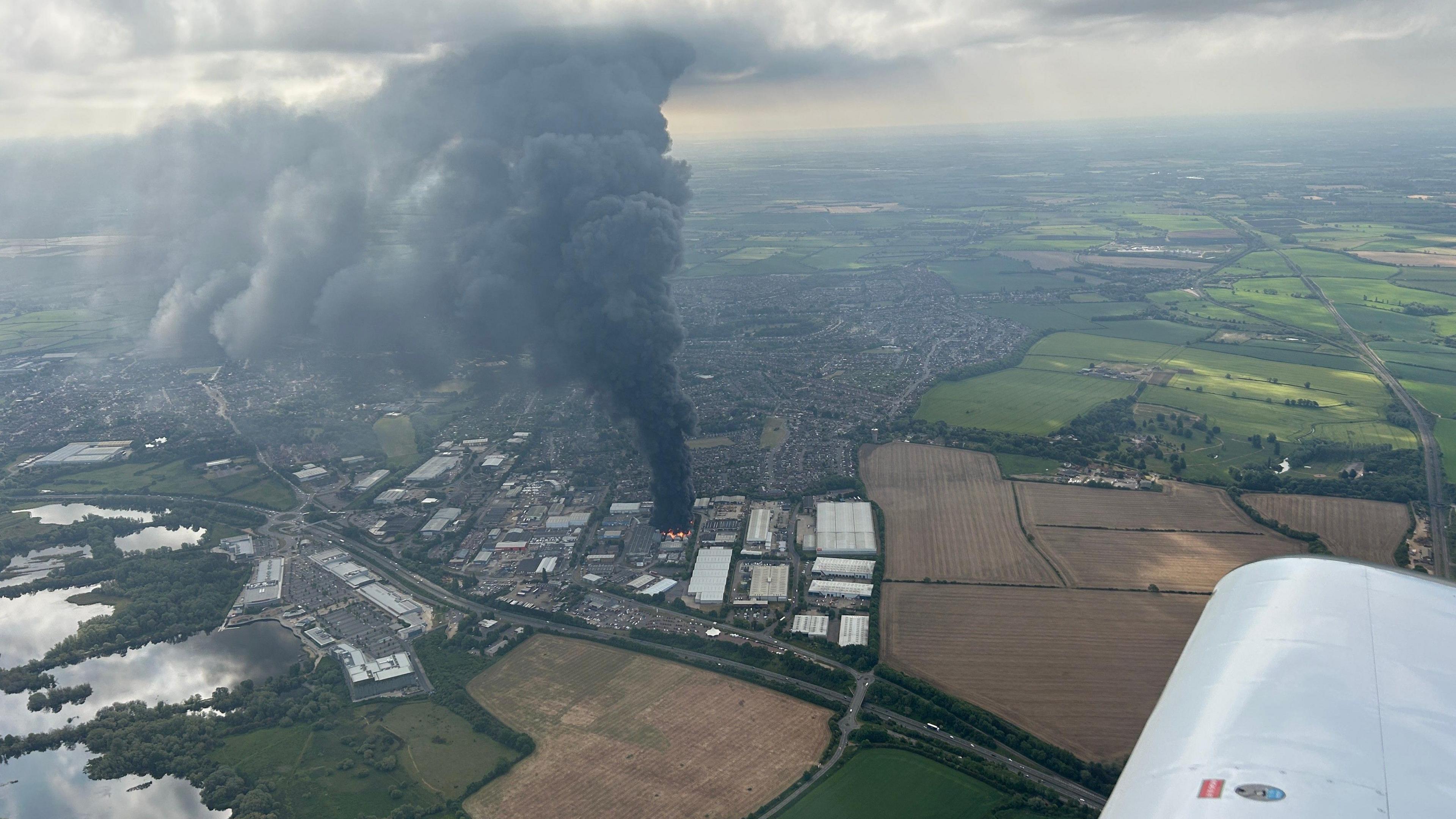 An aerial view of the fire, taken from an airplane, showing a towering plume of black smoke rising into the clouds above. Fields and the nearby town of Rushden are visible surrounding the epicentre of the fire.