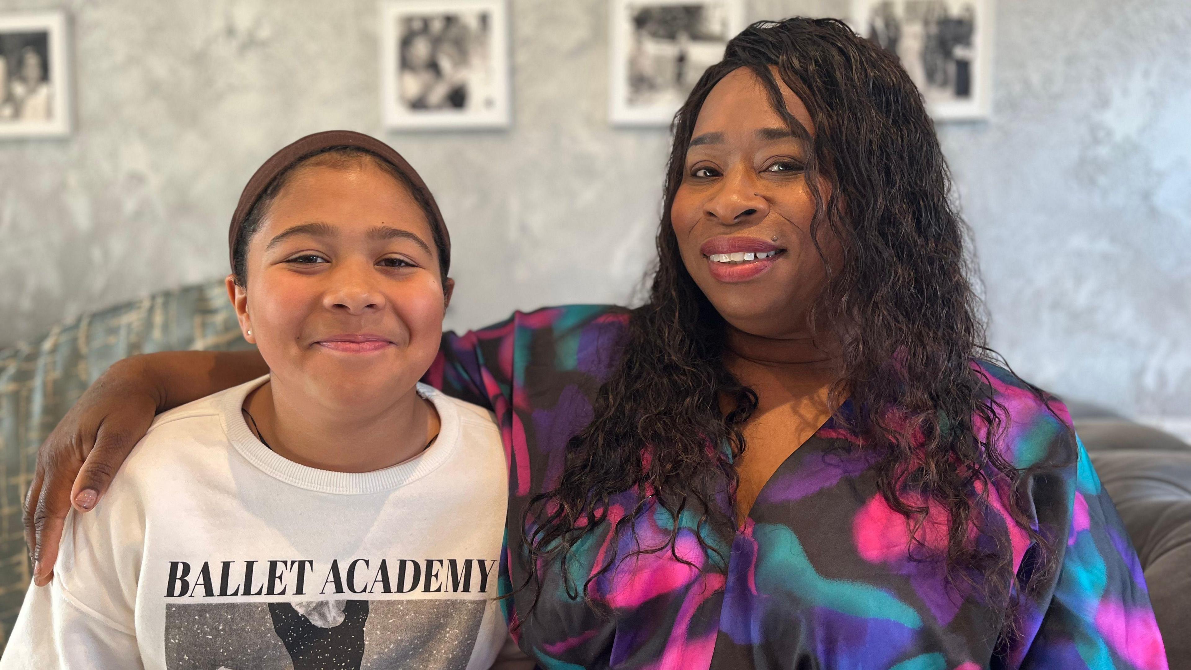 A mum, Dionne, and her daughter Alexis sit on a sofa. Dionne has her arm around Alexis and is wearing a bright pink and black dress. Both are smiling. 