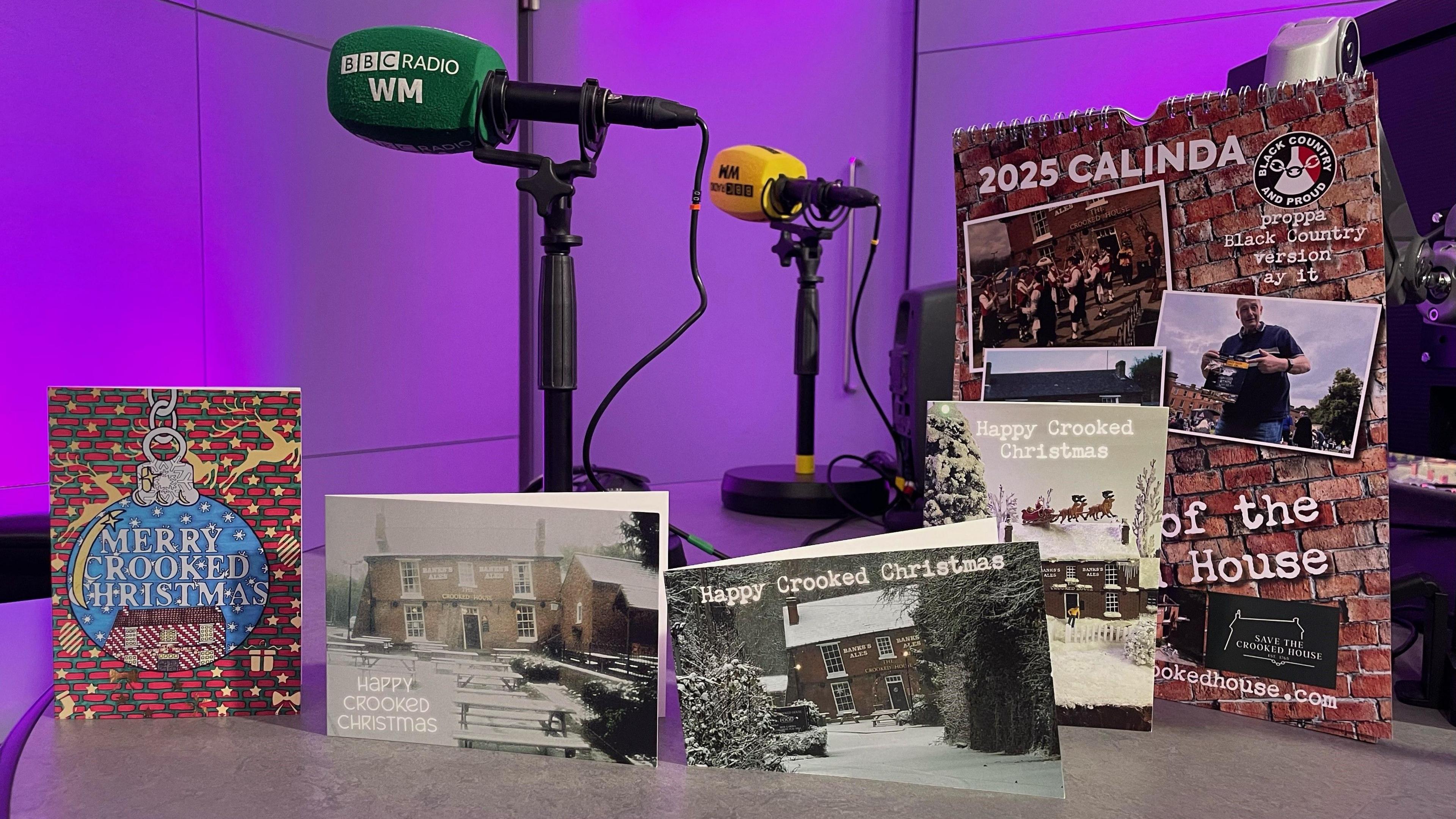 A display of Crooked House Christmas cards and the new calendar placed on a table in the BBC Radio WM studio. The walls behind are purple and two BBC Radio WM microphones are set up behind the display.