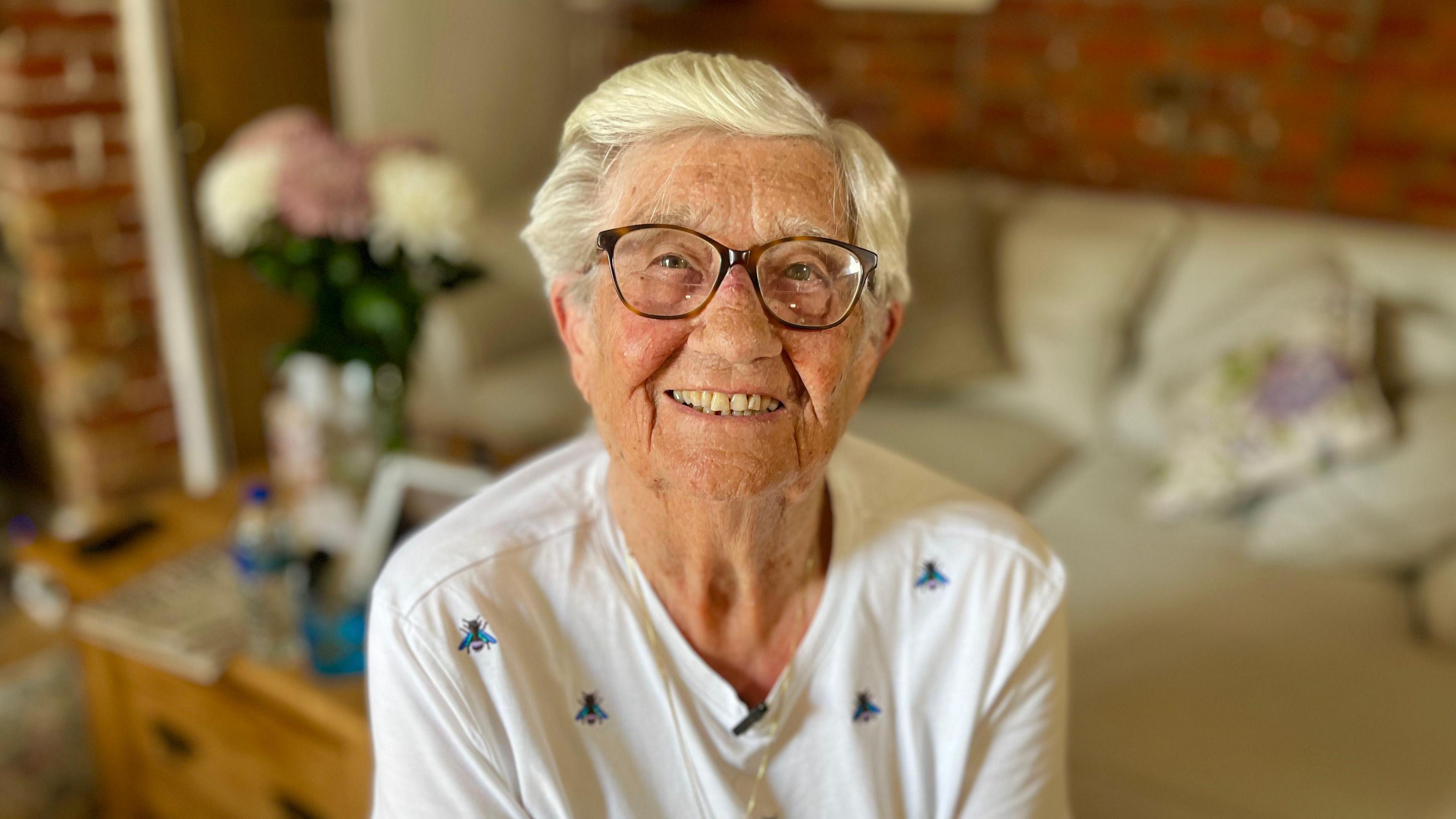 Lady with silver hair and glasses smiles into the camera lens. There are flowers
on a table behind her and a cream sofa with scattered cushions. She is wearing a white top with pictures of bees.